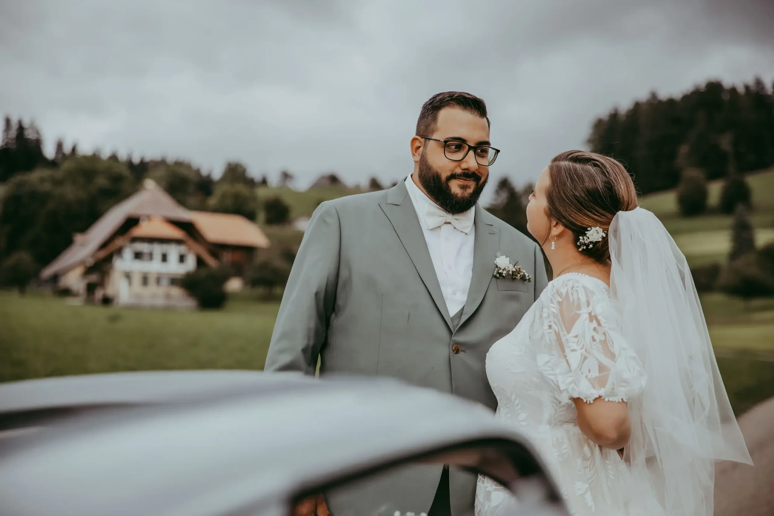 Ein Brautpaar bei einer Hochzeit im Freien, sichtbare Hochzeitskleidung, hinter grüner Landschaft und einem Haus im Hintergrund.