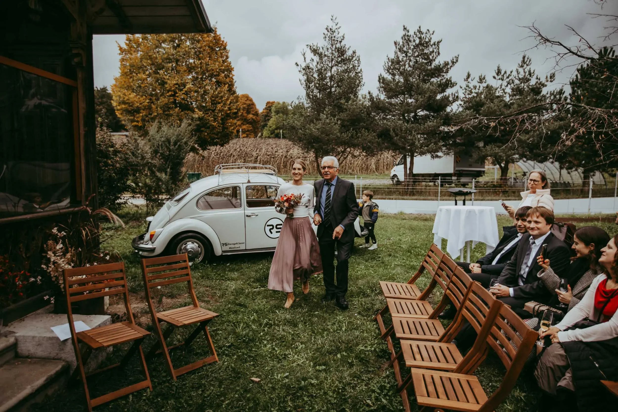 Hochzeit im Garten mit Braut und älterem Mann, Gäste sitzen auf Holzstühlen, ein Oldtimer-VW Käfer im Hintergrund, Herbstbäume und Wolken am Himmel.
