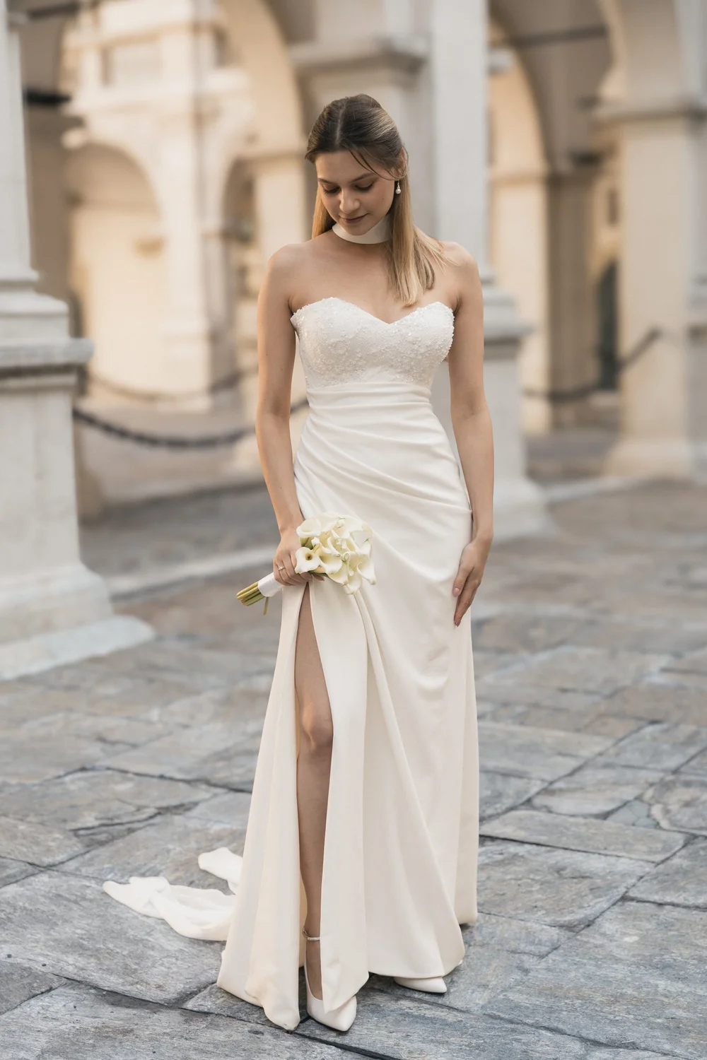 Braut im weißen Kleid mit Calla-Strauß vor den Säulen eines historischen Innenhofs.