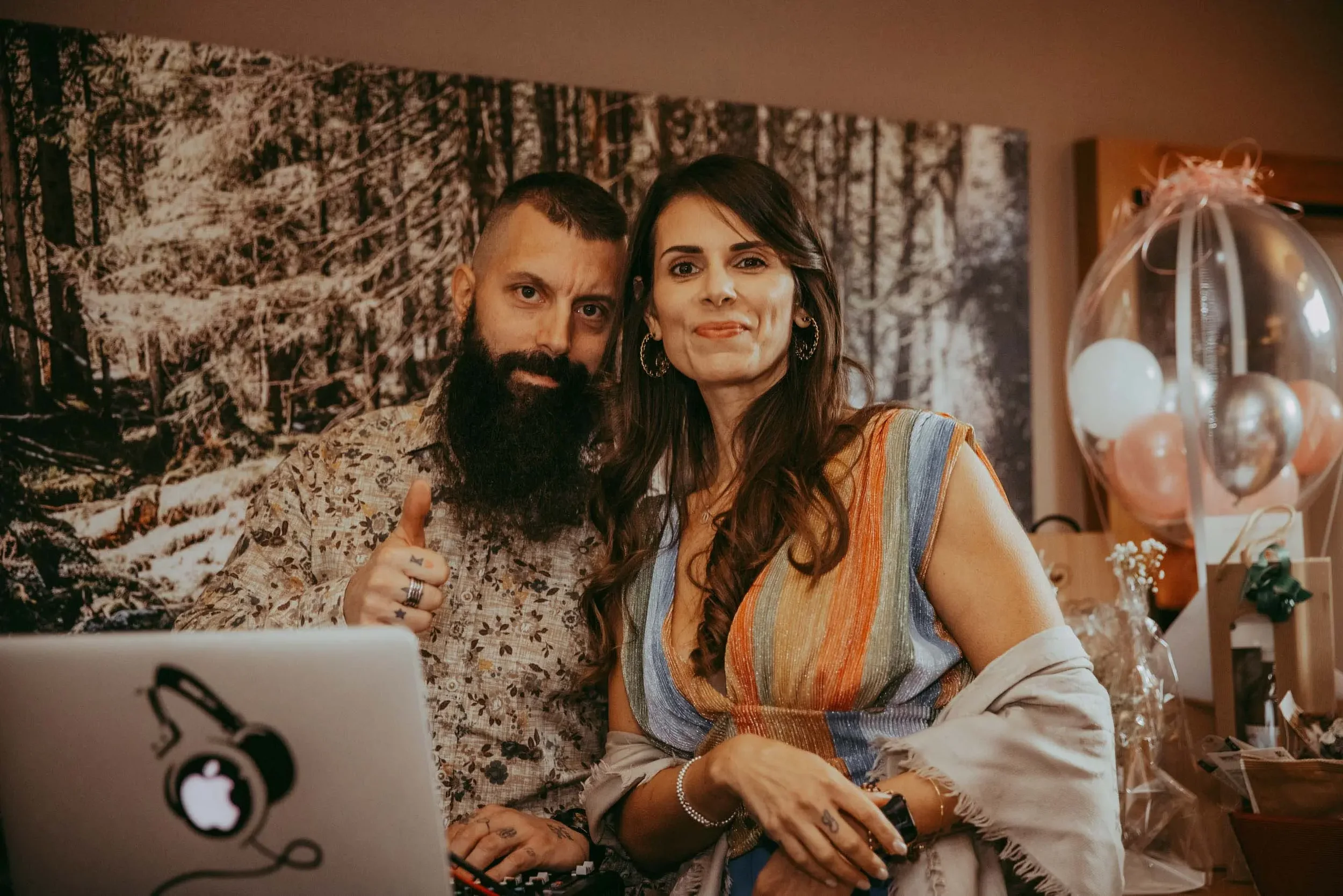Ein Mann mit Bart und eine Frau sitzen vor einem Laptop mit einem Apple-Logo, die im Hintergrund einen Walddruck an der Wand haben. Die Frau trägt ein buntes Kleid, der Mann einen bedruckten Hemd und beide schauen in die Kamera.