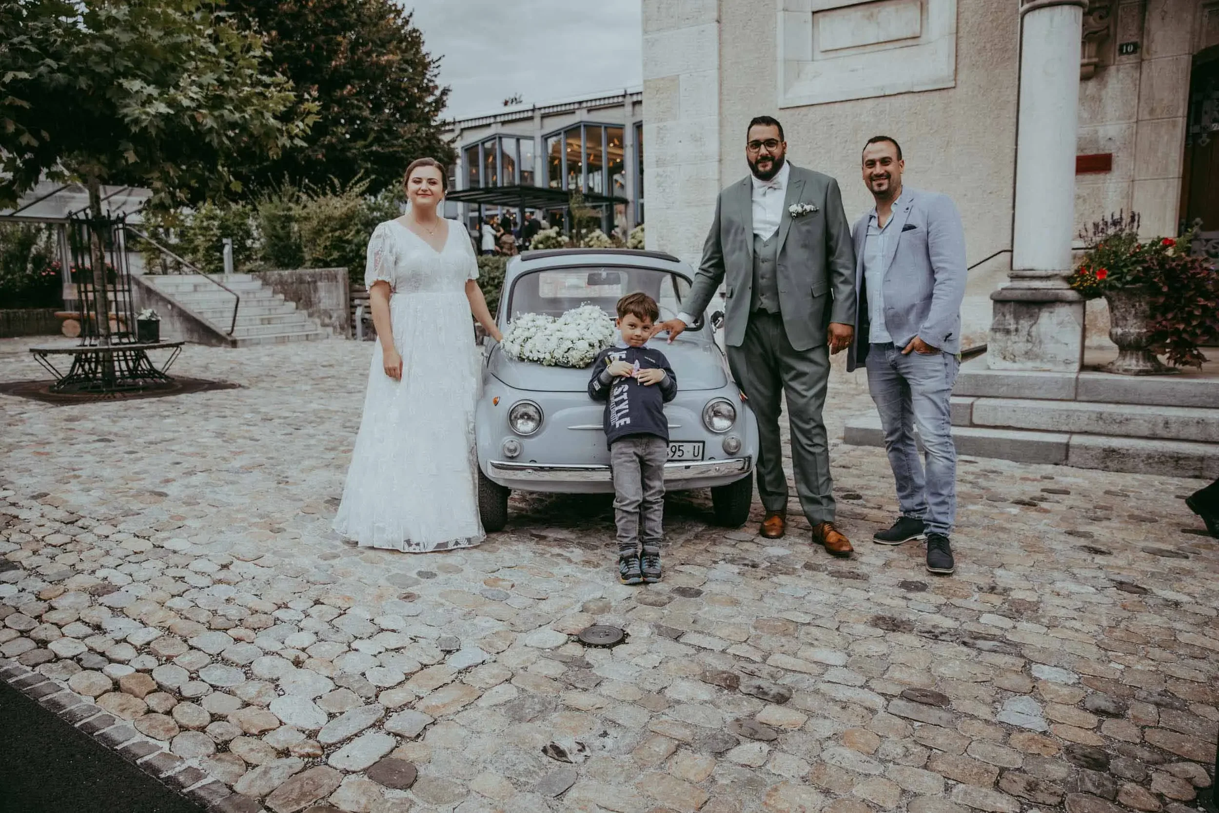 Gruppe von vier Menschen, darunter zwei Männer, eine Frau und ein Kind, die auf einer Steinpflasterstraße vor einem alten Gebäude stehen. Das Kind steht vor einem kleinen Vintage-Auto mit Blumen auf dem Auto. Es ist eine Hochzeitsszene mit einer fest