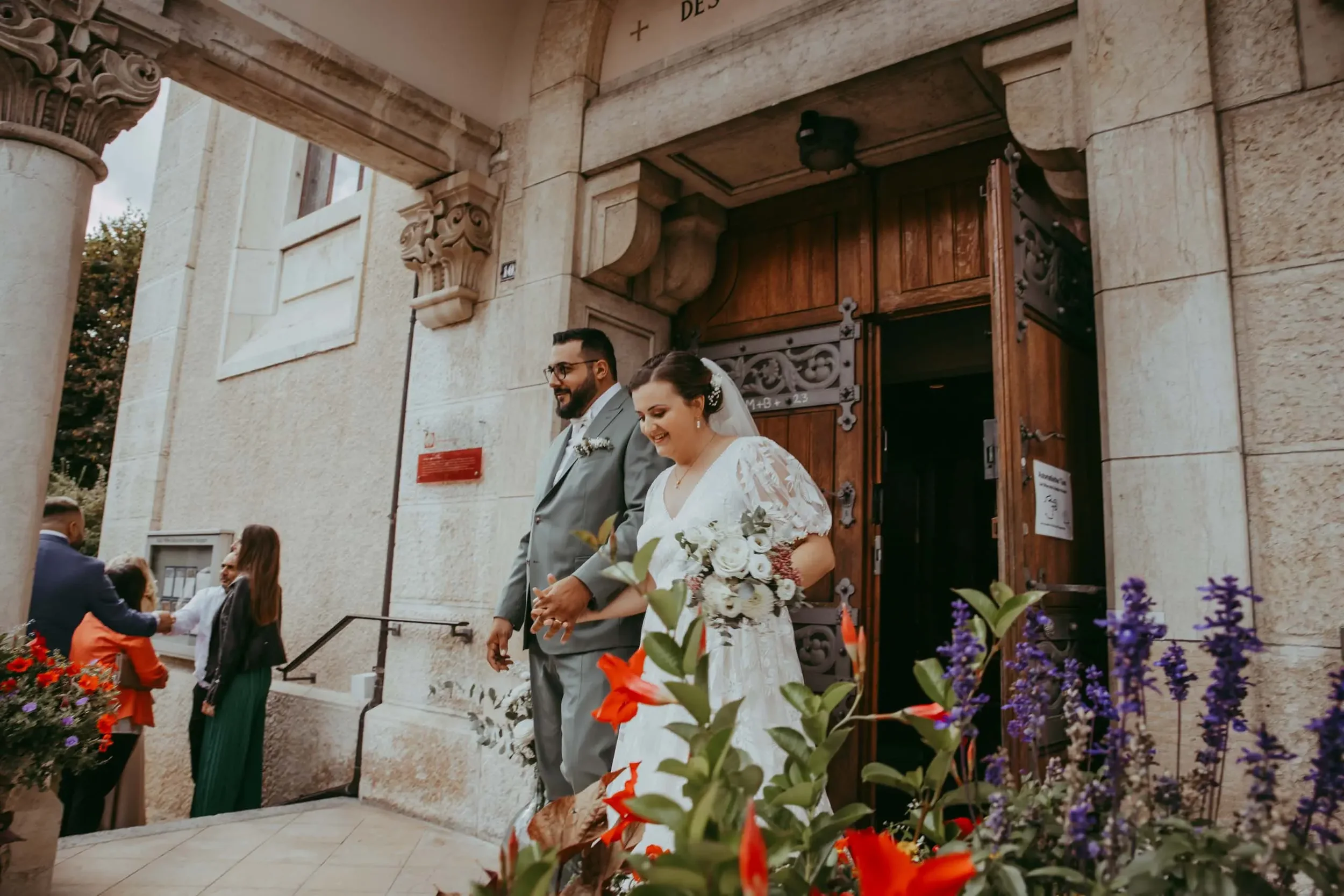 Ein Brautpaar steht vor der Kirche, die Braut trägt ein weißes Kleid mit Blumenmuster und hält einen Blumenstrauß, der Bräutigam trägt einen grauen Anzug und eine Krawatte. Im Vordergrund sind bunte Blumen zu sehen, im Hintergrund weitere Gäste vor d
