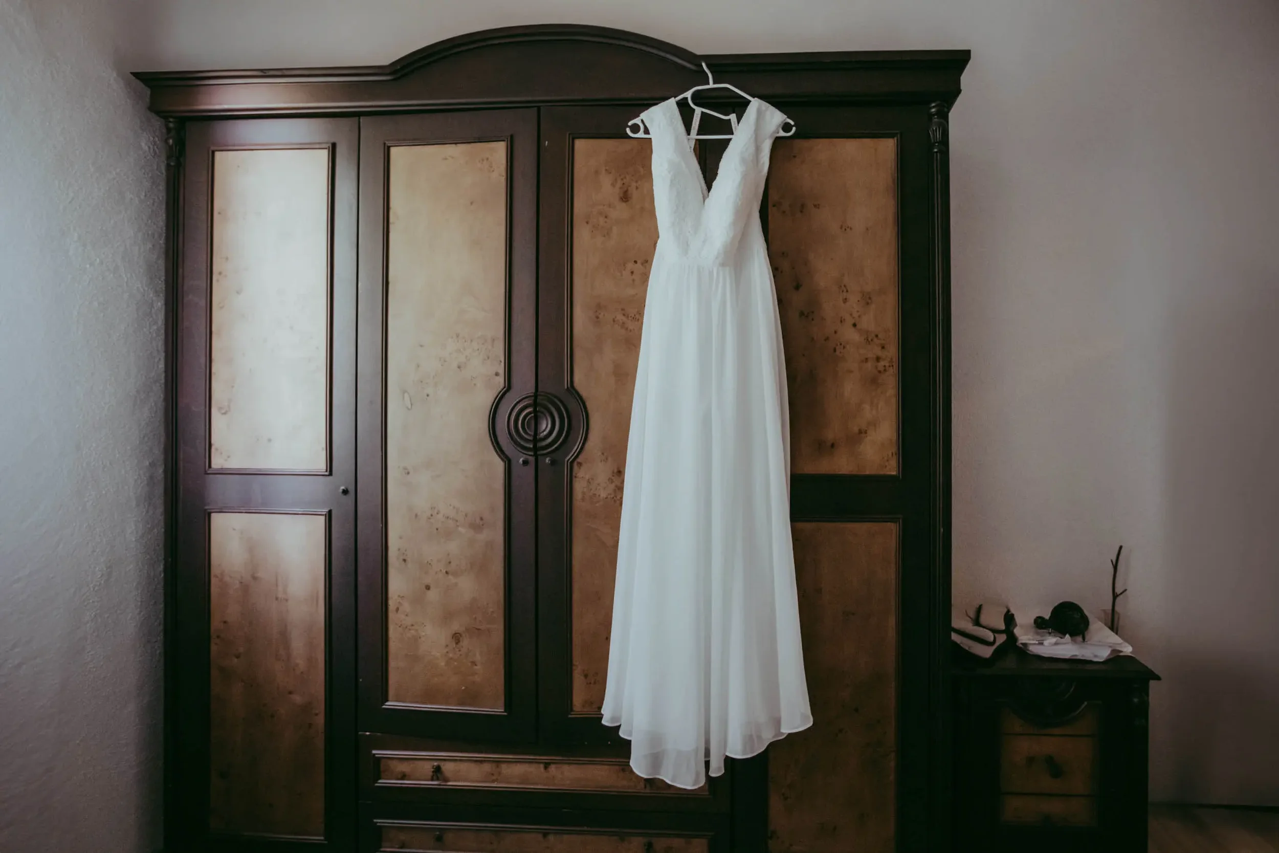 Ein weißes Brautkleid hängt an einem weißen Kleiderbügel an einem dunklen Kleiderschrank in einem Raum.