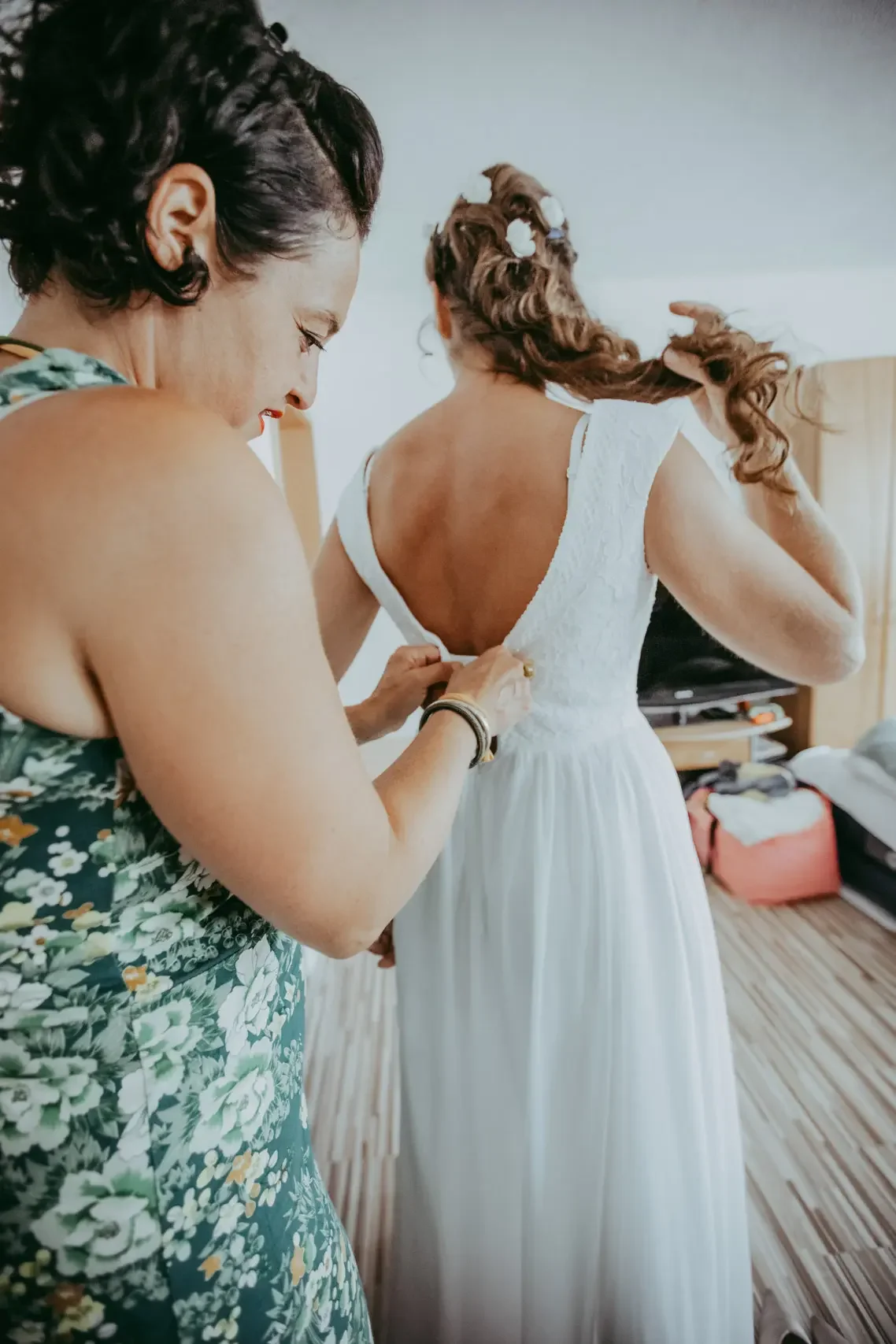 Eine Frau hilft einer Braut, das Brautkleid anzuprobieren, im Zimmer mit Holzfußboden und Koffer.