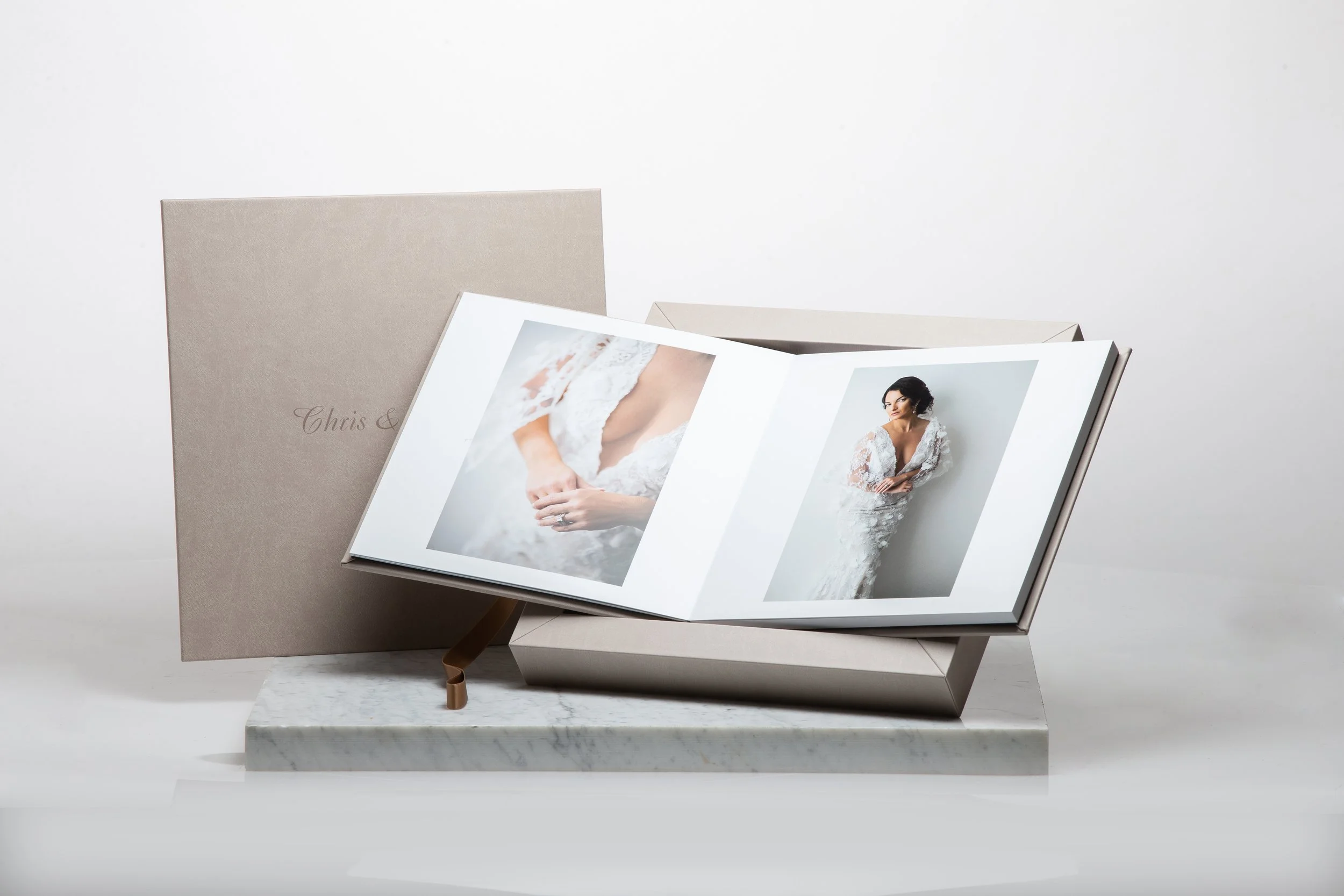 Hochzeitsfotobuch auf einem weißen Tisch, beige Tasche im Hintergrund, offene Seite zeigt Fotos einer Braut in Weiß. Moderne, elegante Präsentation.