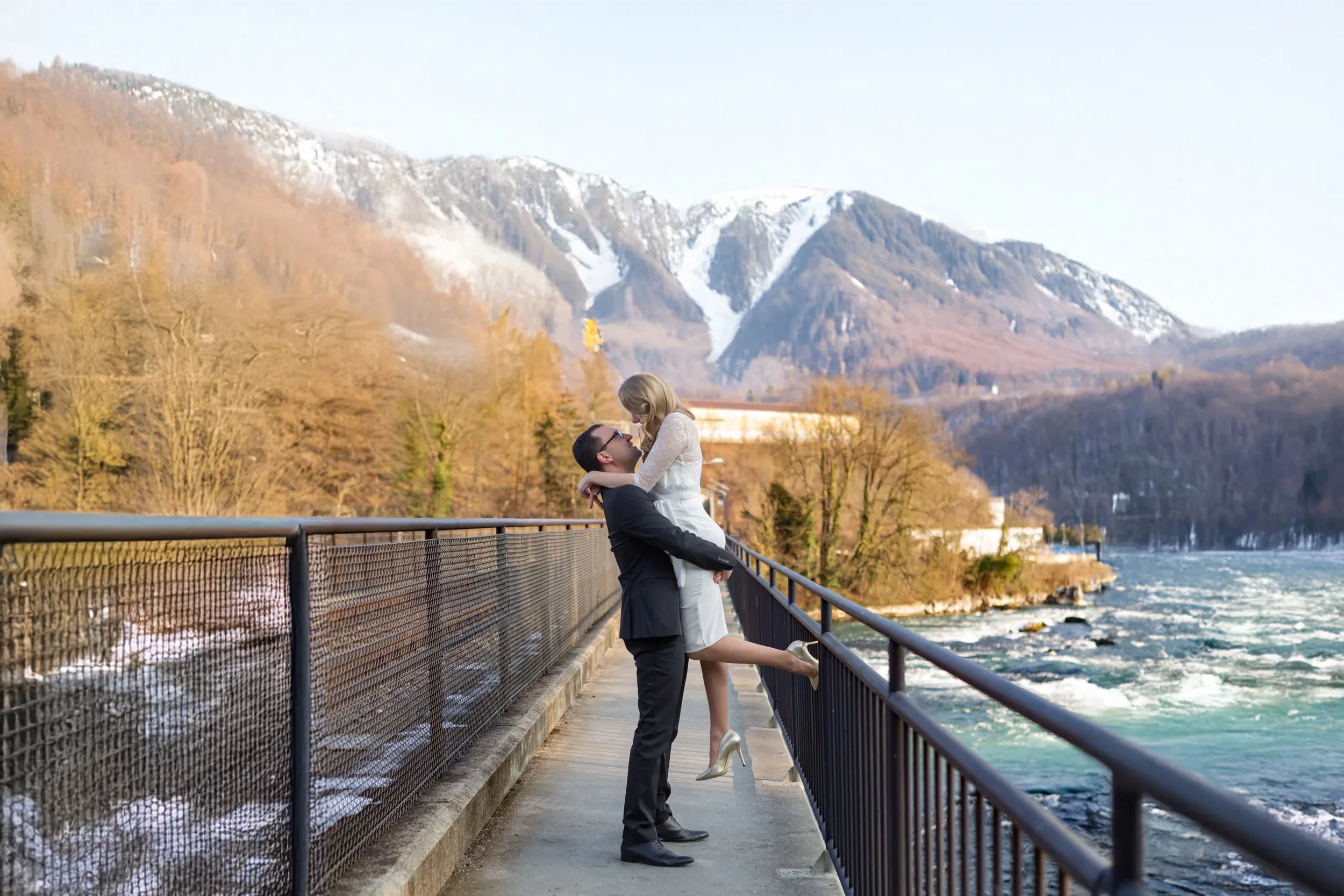 Ein Mann hebt eine Frau auf dem Gesicht, beim Blick auf einen Fluss mit Bergen im Hintergrund, während sie sich anlächelt.