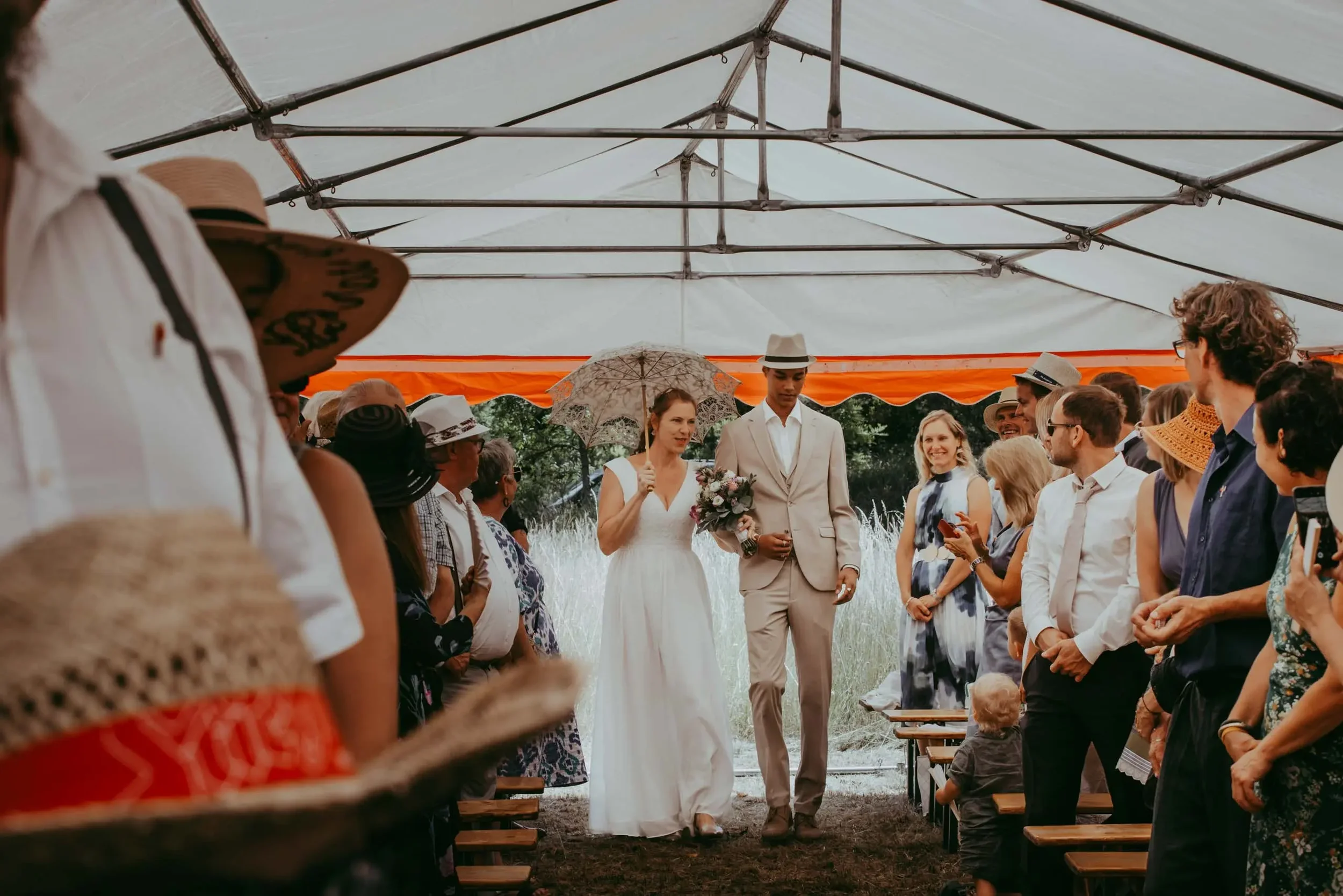Ein Brautpaar während ihrer Hochzeit, umgeben von Gästen unter einem Zelt, bei gutem Wetter.