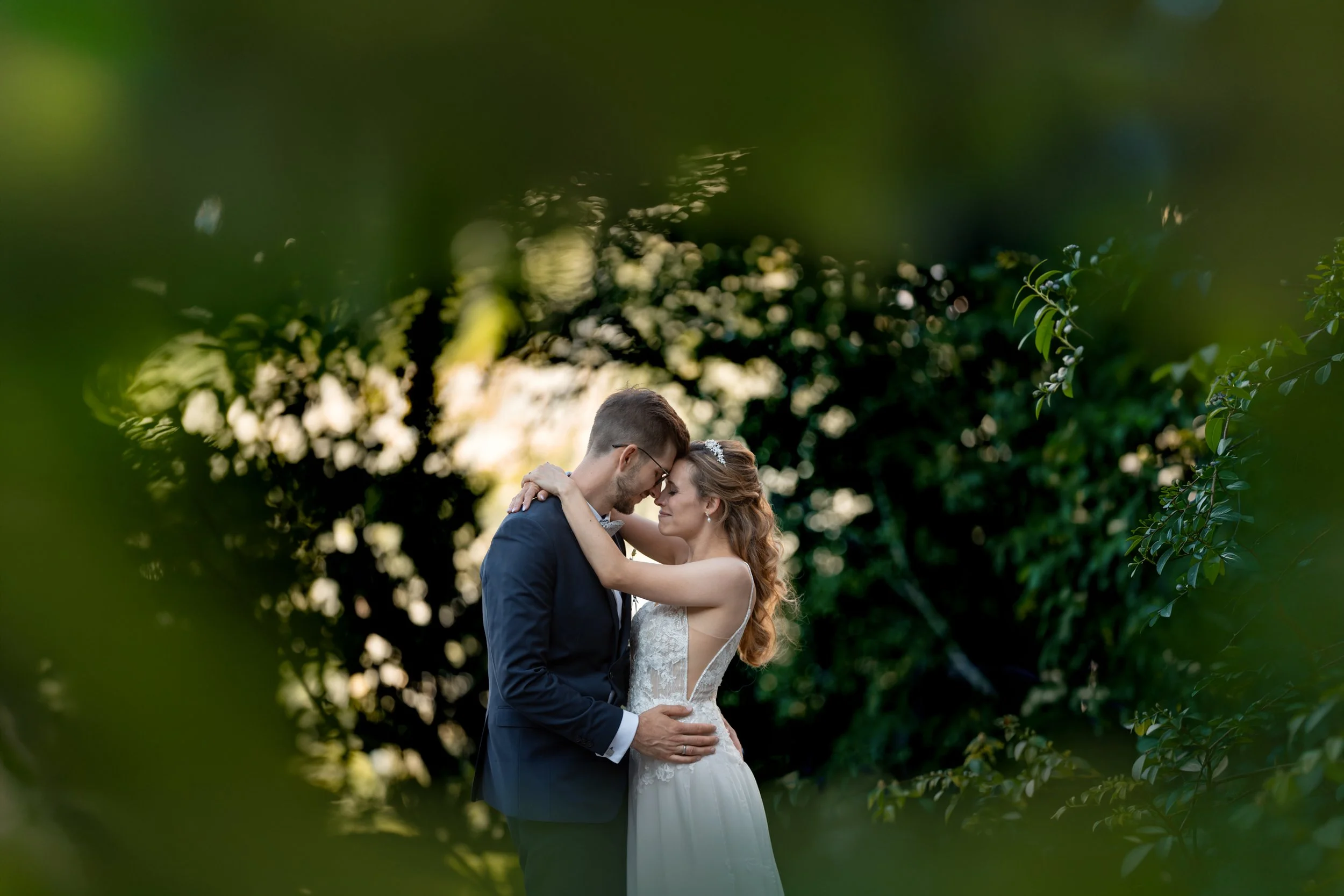 Ein heiratendes Paar umarmt sich in einer grünen, bewaldeten Umgebung bei Sonnenuntergang.