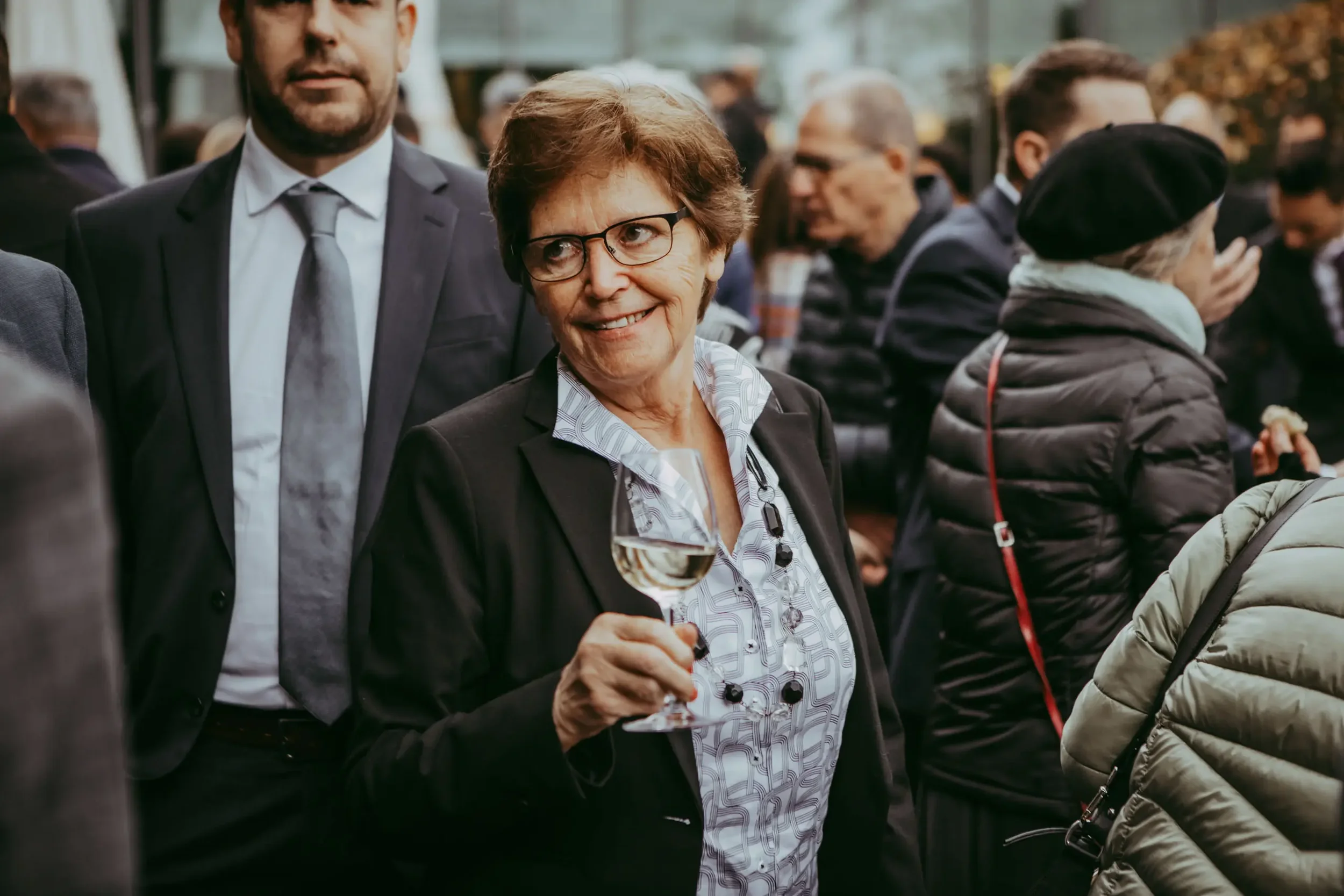 Eine ältere Frau trägt eine Brille, hält ein Weinglas und lächelt bei einer gesellschaftlichen Veranstaltung im Freien, umgeben von anderen Menschen, die sich unterhalten.