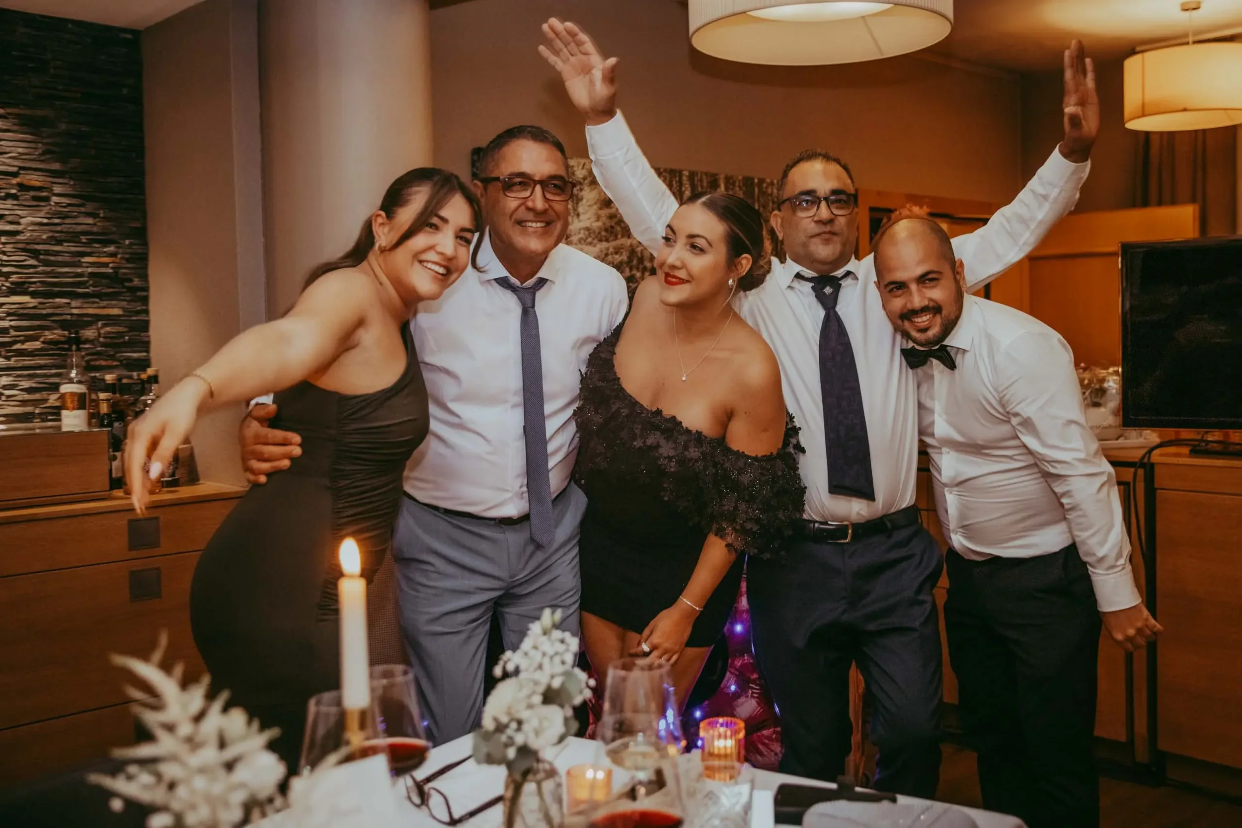 Eine Gruppe von sechs Menschen, die auf einer Feier zusammenstehen und lächeln. Es sind drei Männer und drei Frauen, die in einem festlichen Raum gekleidet sind. Im Vordergrund sind Kerzen und eine Blumenarrangement auf einem Tisch zu sehen.