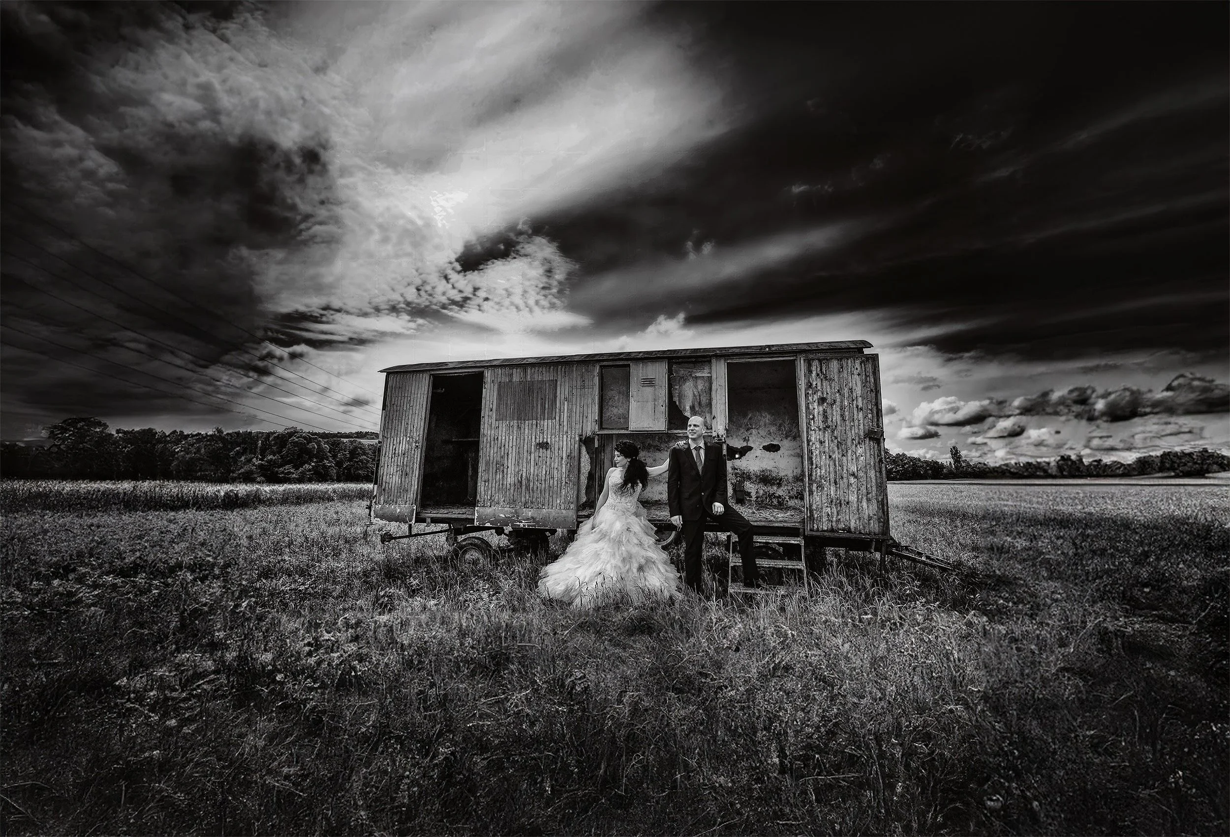 Ein schwarz-weiß Bild von einem Paar in formeller Kleidung, das neben einem alten, offenen Holzwohnwagen in einem Feld steht, mit dunklem, dramatischem Himmel im Hintergrund.