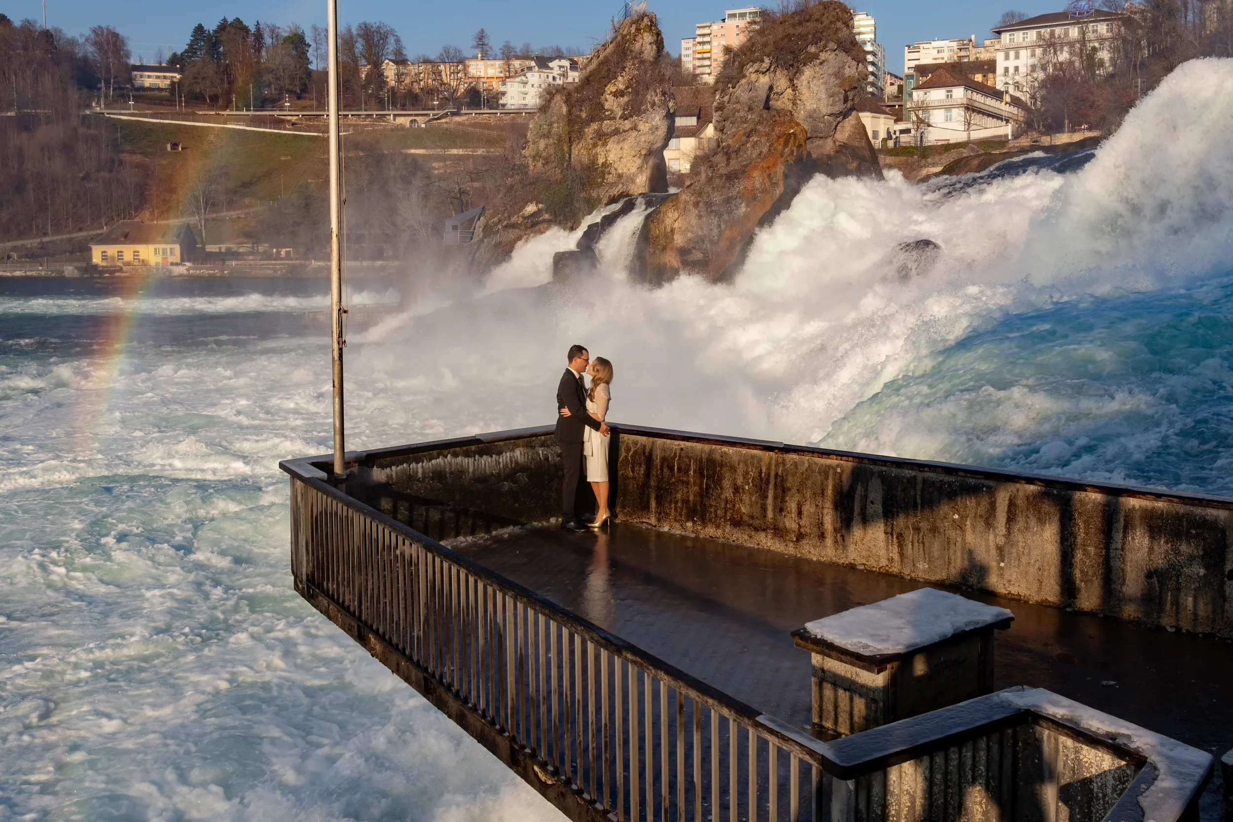 Ein verliebtes Paar steht auf einem Wasserbalkon vor einem Wasserfall, hält sich an den Händen, umarmt sich liebevoll, im Hintergrund sind Häuser und Bäume, bei Sonnenuntergang, mit einem Regenbogen im Dunst.