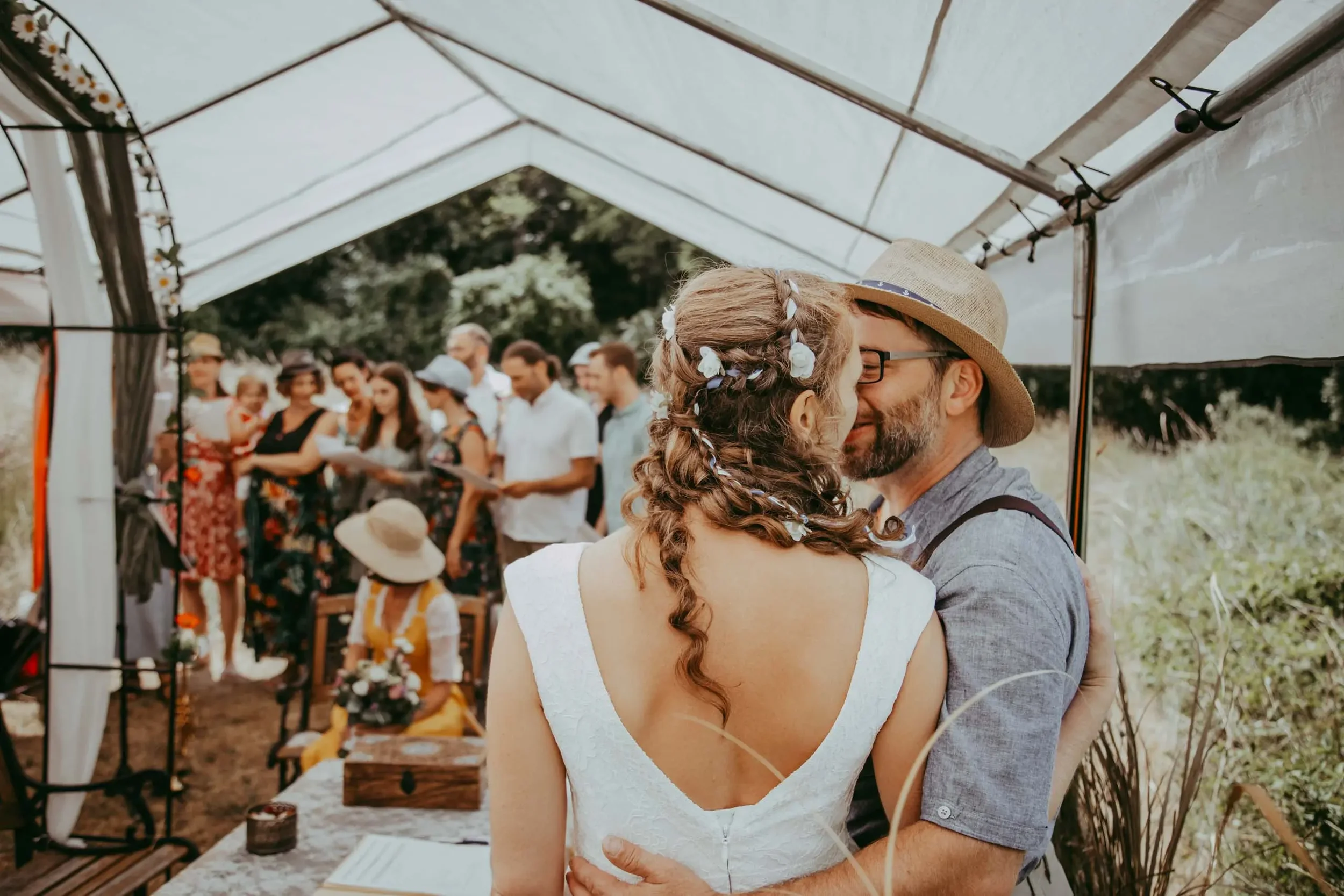 Ein verliebtes Paar umarmt sich unter einem Zelt bei einer Hochzeit im Freien, im Hintergrund sind Gäste zu sehen.