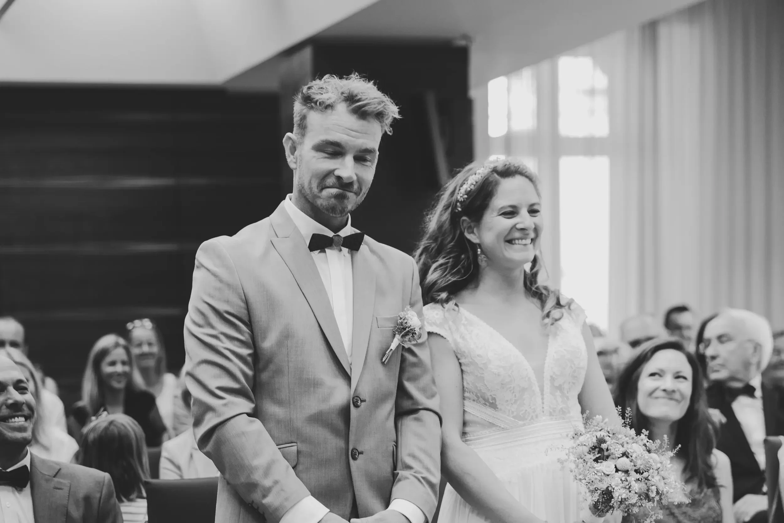 Ein Brautpaar steht während einer Trauungszeremonie in einem Raum mit großen Fenstern. Es ist schwarz-weiß aufgenommen. Die Frau trägt ein Hochzeitskleid und hält einen Blumenstrauß, während der Mann einen Anzug mit Fliege trägt.