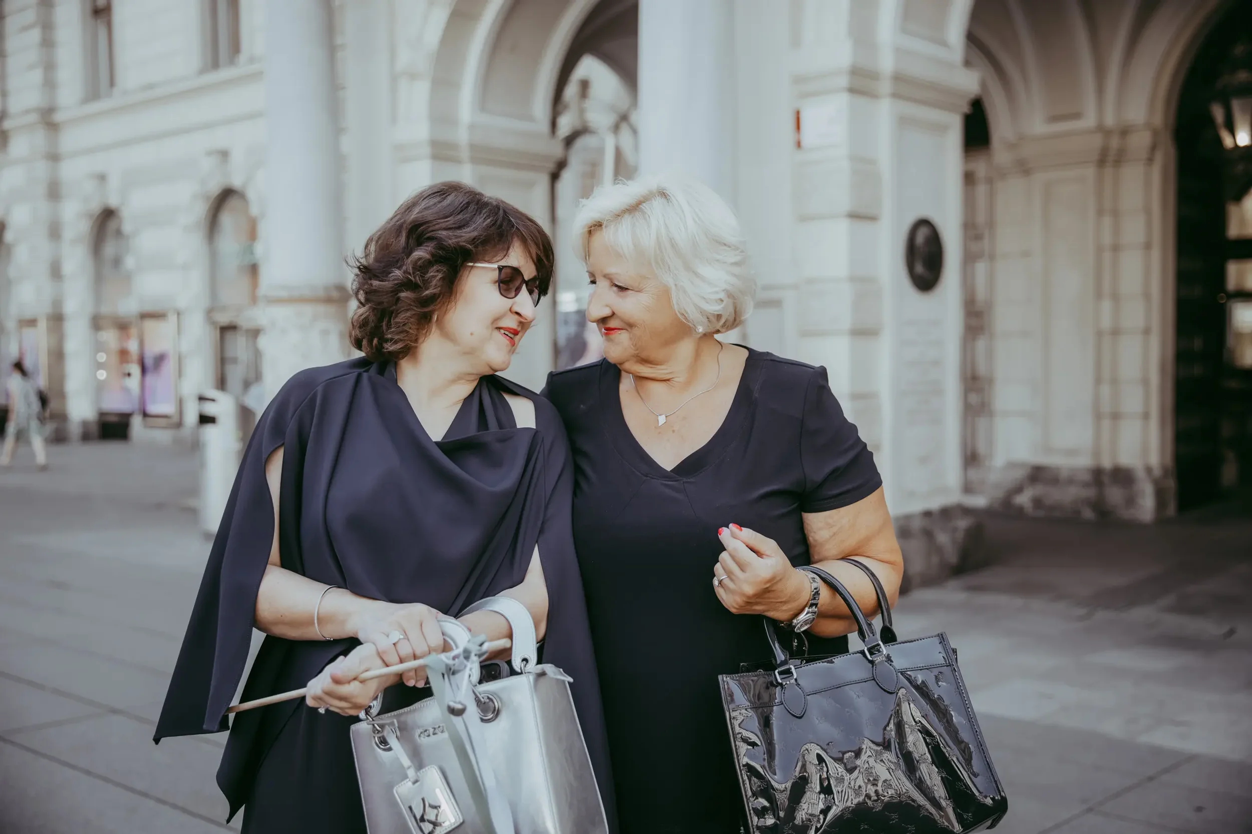 Zwei Frauen, eine ältere und eine jüngere, teilen einen Moment der Freude, beide lächelnd und sich berührend, vor einem historischen Gebäude. Die ältere Frau hat kurze weiße Haare und trägt eine schwarze Bluse, die jüngere Frau hat braune Haare, träg
