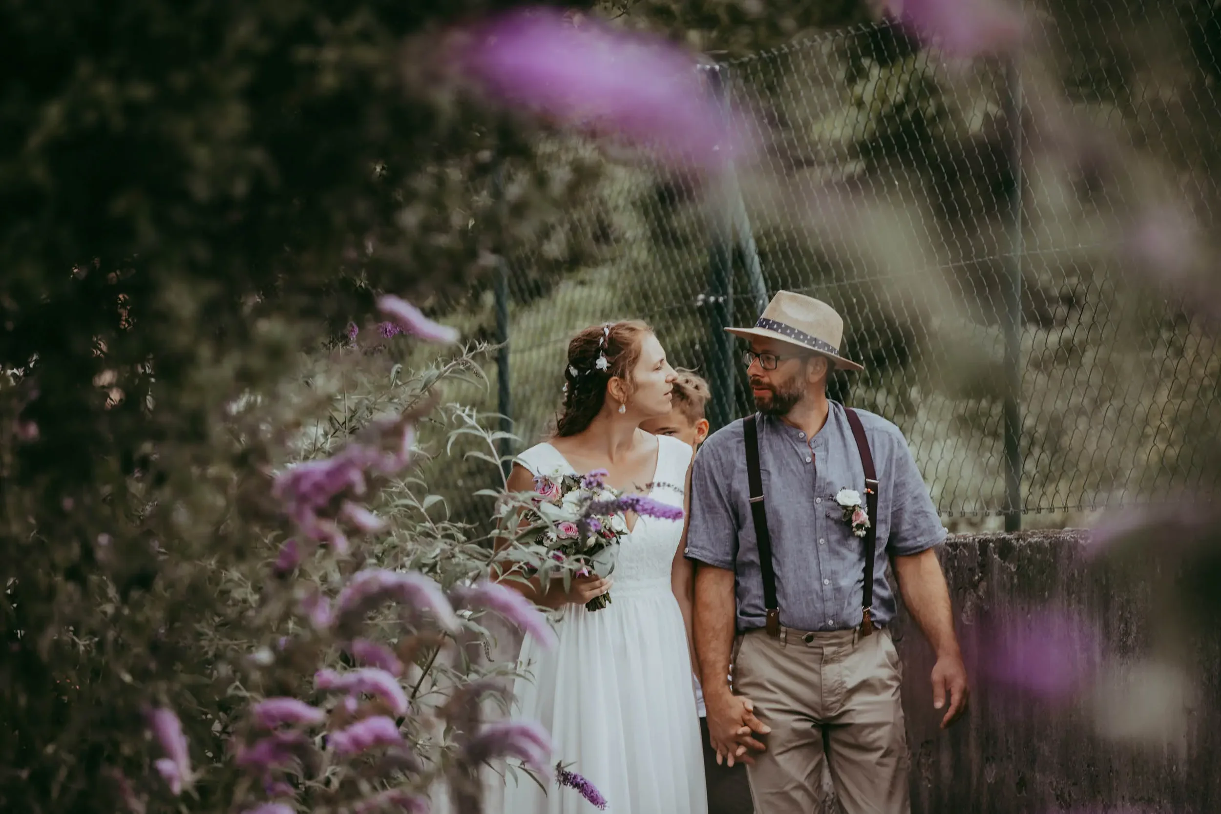 Ein Brautpaar mit einem Kind, die Händchen haltend, in einem Garten mit Blumen, im Gespräch, bei einer Hochzeit.