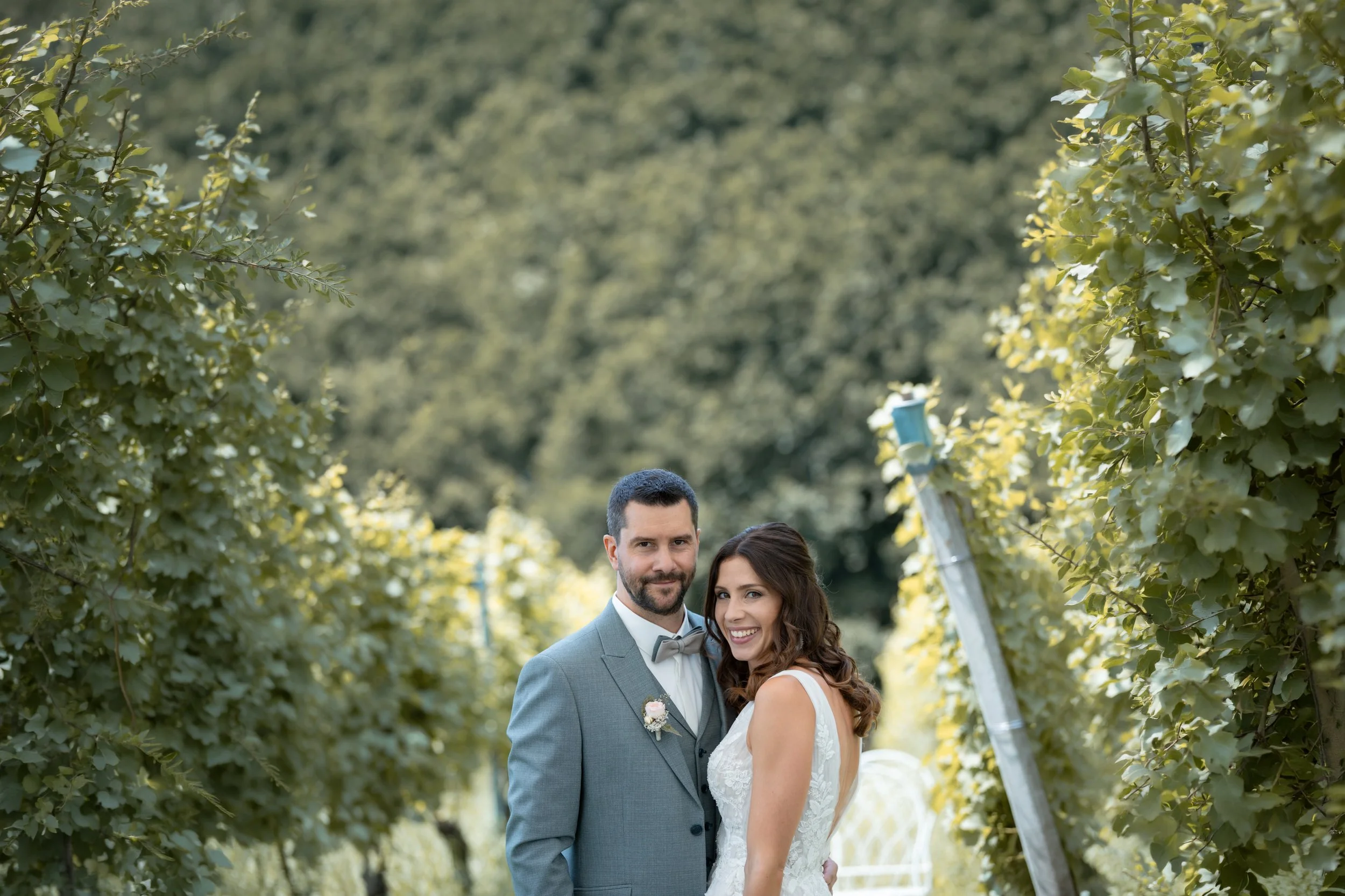 Ein Brautpaar steht in einem Garten zwischen grünem Laub, vermutlich an ihrem Hochzeitstag.