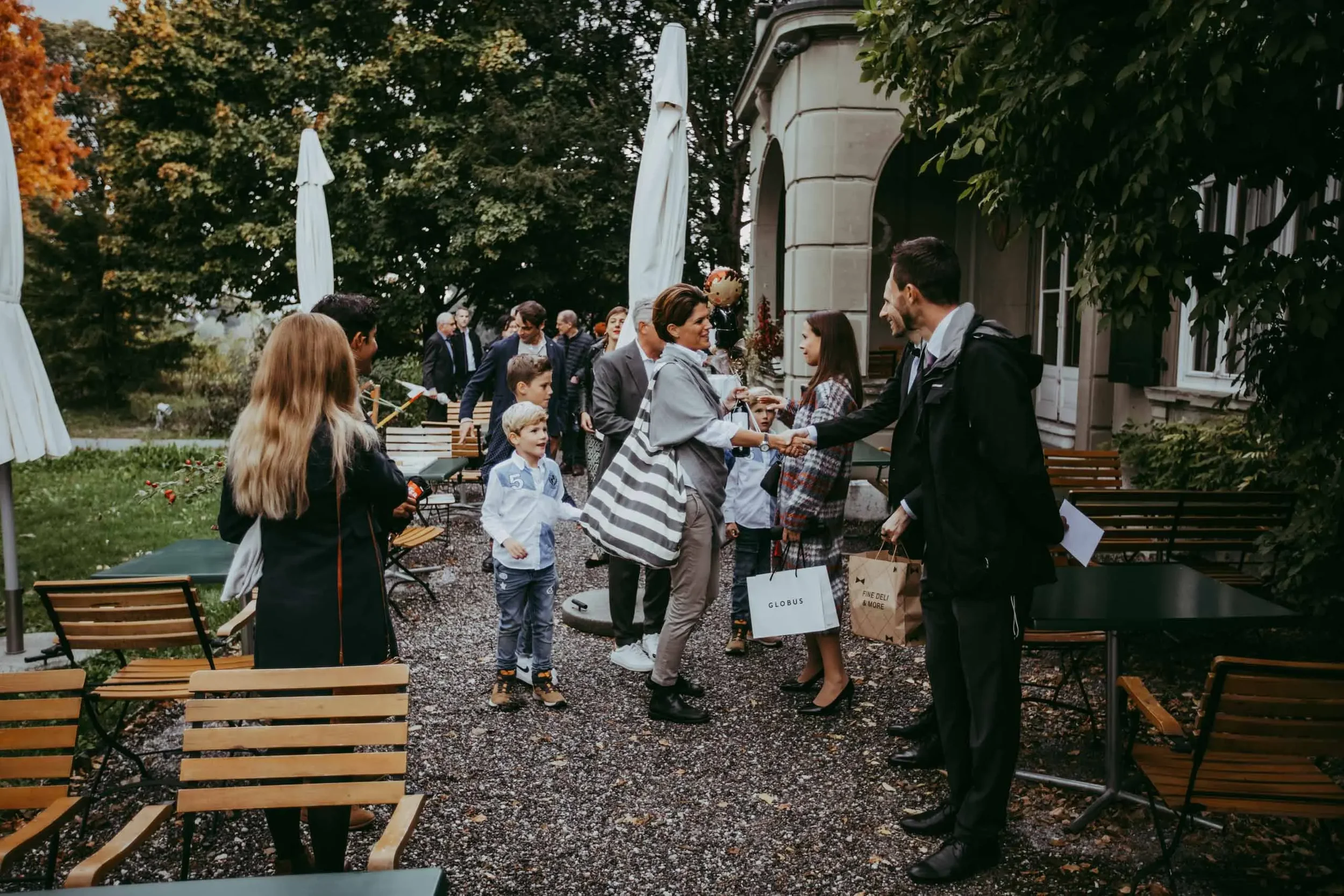 Menschen begrüßen sich auf einer Terrasse vor einem großen Gebäude im Freien, umgeben von Bäumen, mit Tischen und Stühlen, einige in Gespräche vertieft, mehrere Handtaschen und Einkaufstüten sichtbar.