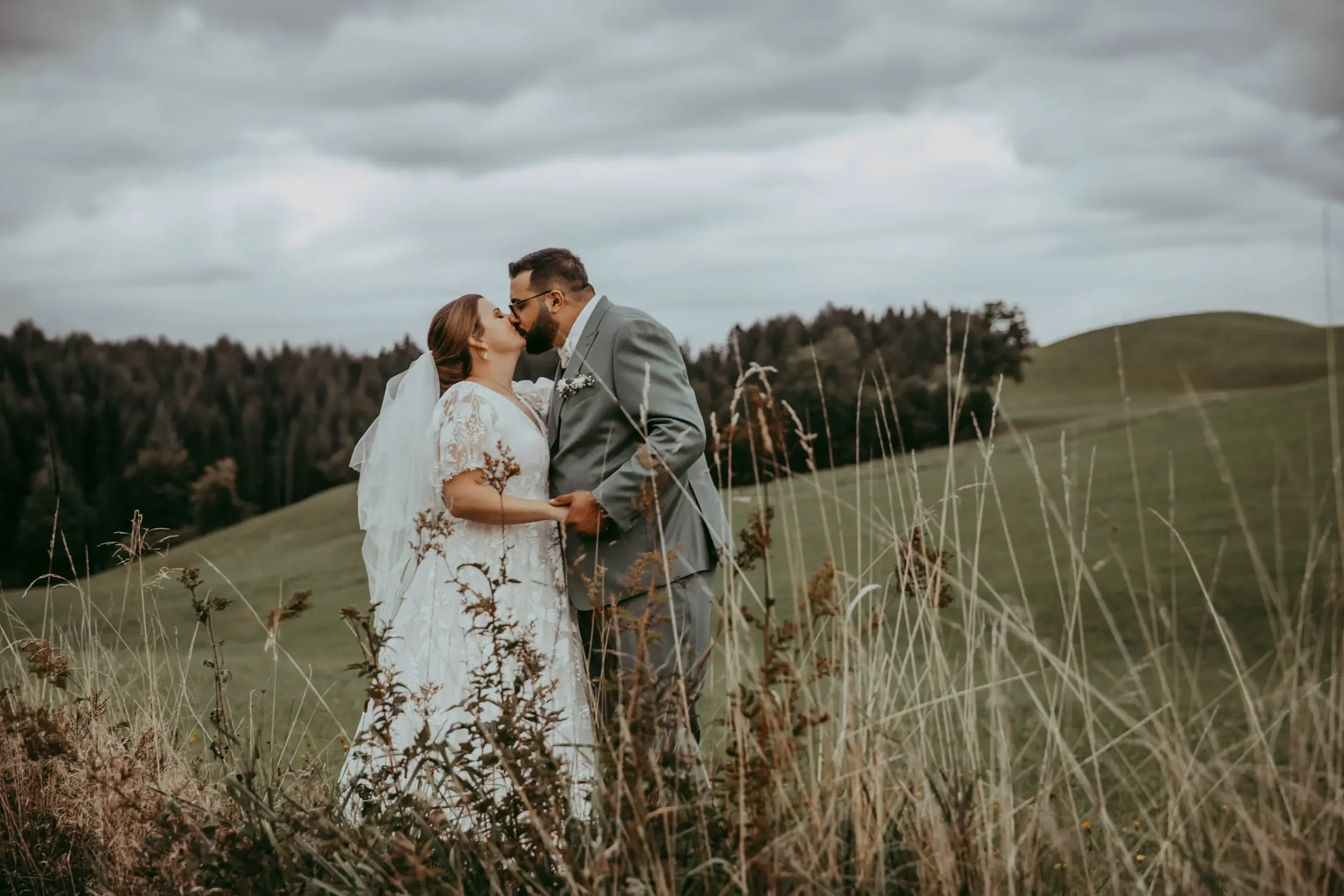 Ein Hochzeitsbild eines Paars, das sich in einem Feld bei bewölktem Himmel küsst. Er trägt einen grauen Anzug, sie ein weißes Brautkleid mit Schleier.