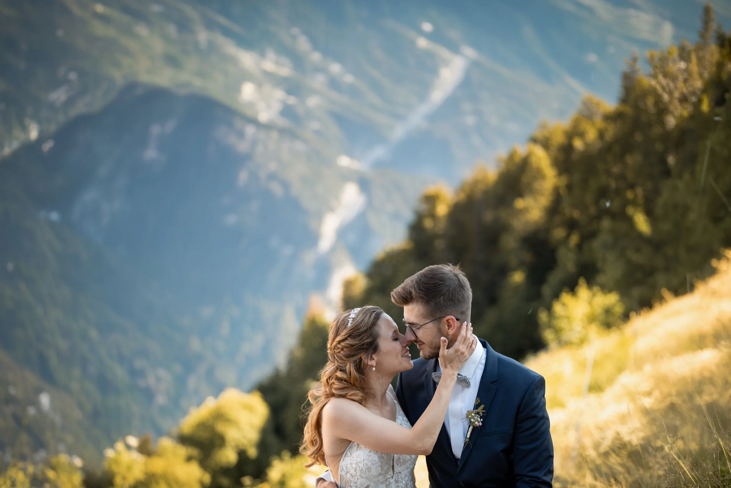 Ein junges Paar, ein Mann und eine Frau, stehen sich nahe und schauen sich liebevoll an, in einer malerischen Berglandschaft im Freien bei Sonnenuntergang. Die Frau trägt ein weißes Brautkleid mit Spitze und ein Tiara, der Mann trägt einen dunklen An