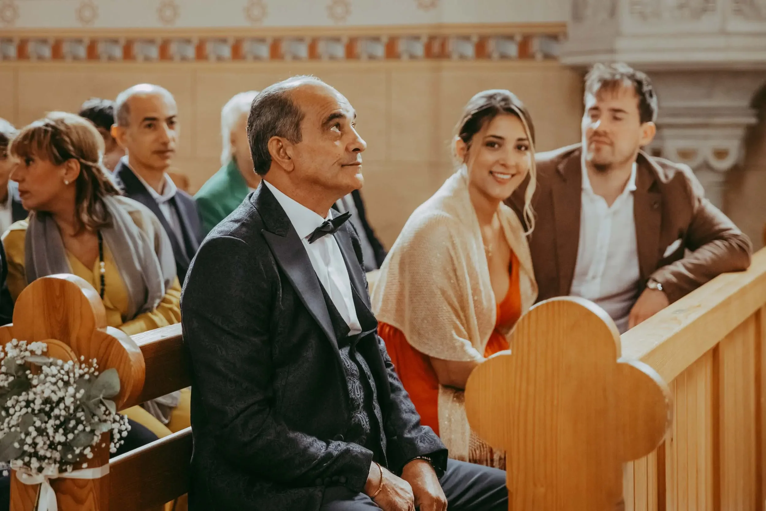 Menschen sitzen in einer Kirche bei einer Zeremonie. Der Fokus liegt auf einem älteren Mann in Smoking und Krawatte, daneben eine lächelnde junge Frau in orangefarbenem Kleid und eine junge Man in brauner Jacke. Im Hintergrund weitere Personen.