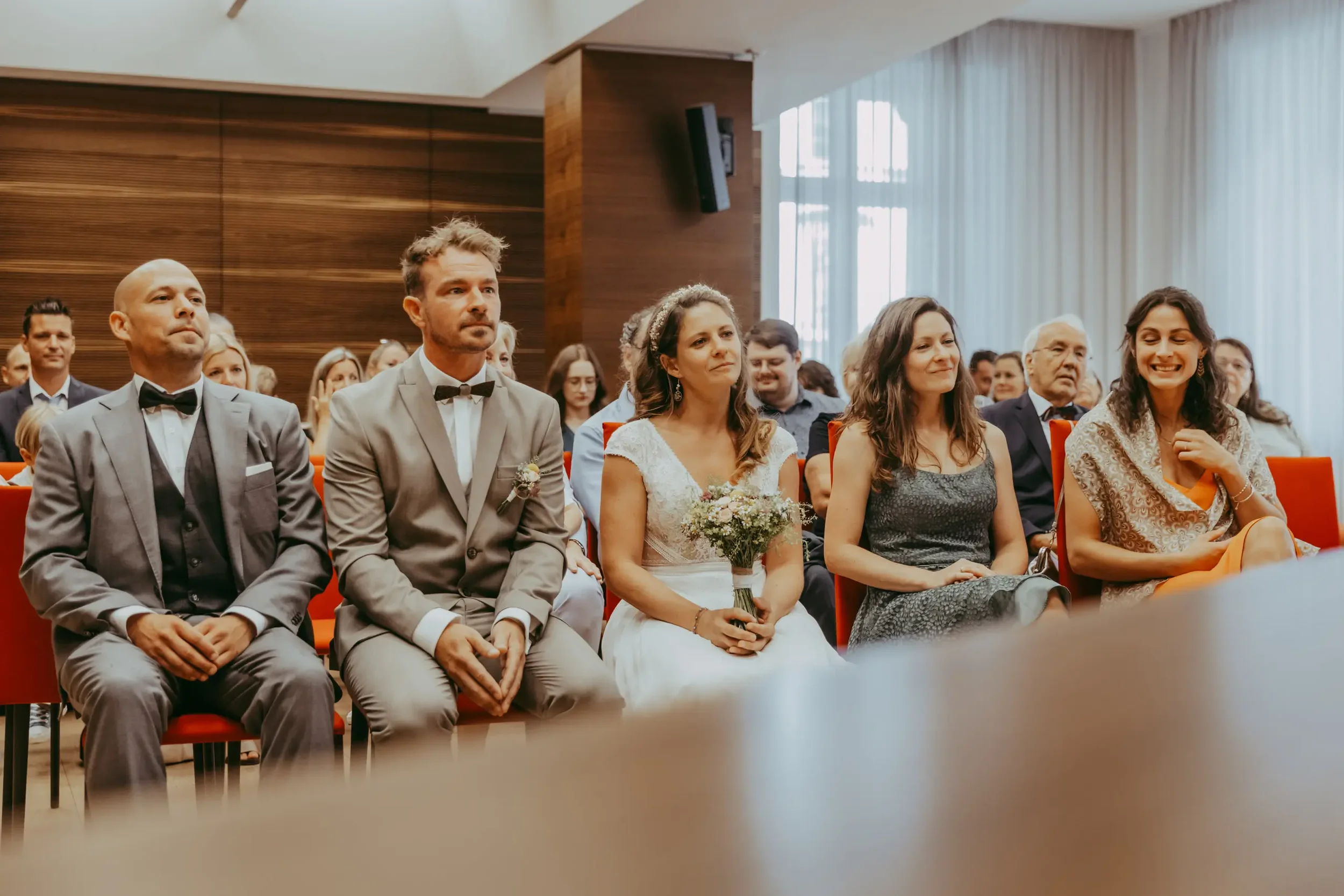 Menschen bei einer Hochzeitsfeier im Saal, einige tragen elegante Kleidung, darunter ein Brautpaar in der ersten Reihe, die Braut hält einen Blumenstrauß.
