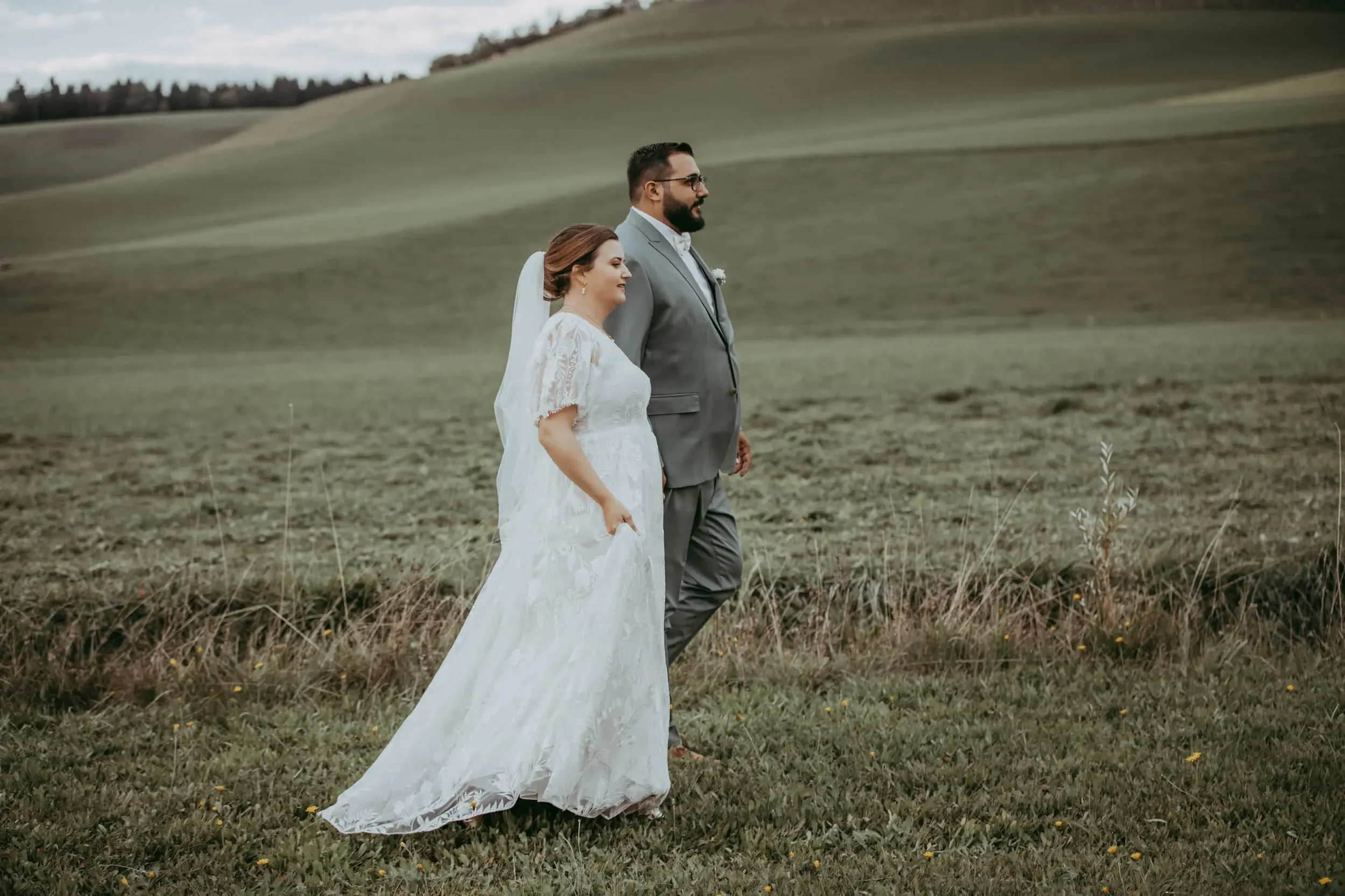 Ein Brautpaar, eine Frau in einem weißen Brautkleid und ein Mann in einem grauen Anzug, läuft Hand in Hand durch eine offene Wiesenlandschaft mit Hügeln im Hintergrund.