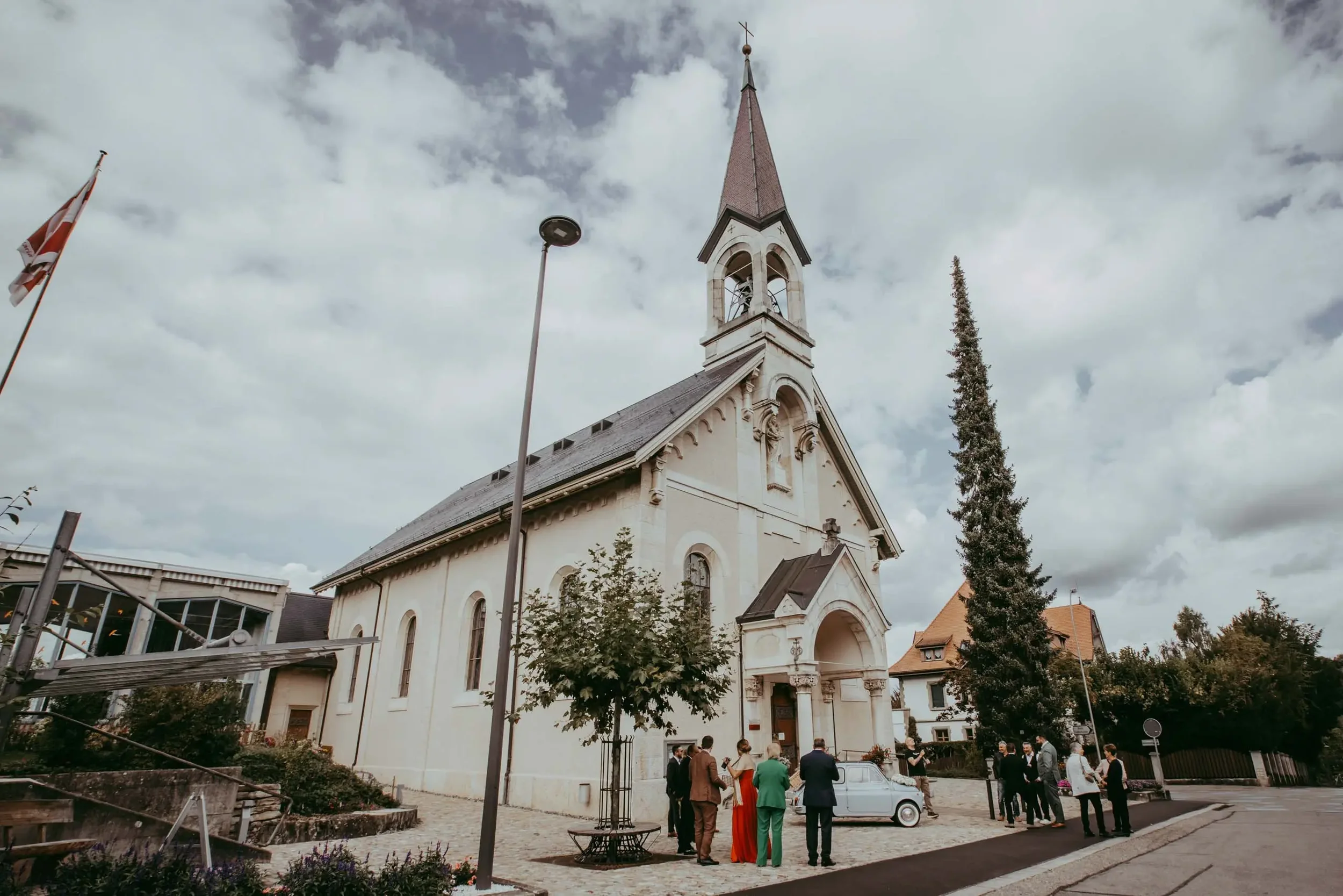 Eine weiße Kirche mit einem hohen schmalen Turm und einem Kreuz oben. Es versammeln sich mehrere Menschen vor der Kirche, einige tragen formelle Kleidung. Der Himmel ist bewölkt und im Hintergrund sind Bäume und Gebäude zu sehen.