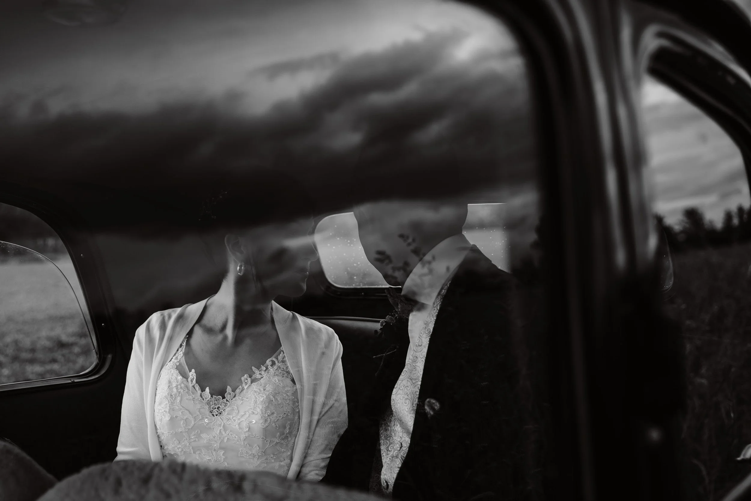 Ein Brautpaar sitzt im Auto, sichtbar durch die Autofenster, die Frau trägt ein weißes Brautkleid mit Spitze und der Mann trägt einen Anzug. Es ist ein schwarzer und weißer Fotostil, der das Paar in einem Intimitätsmoment zeigt.