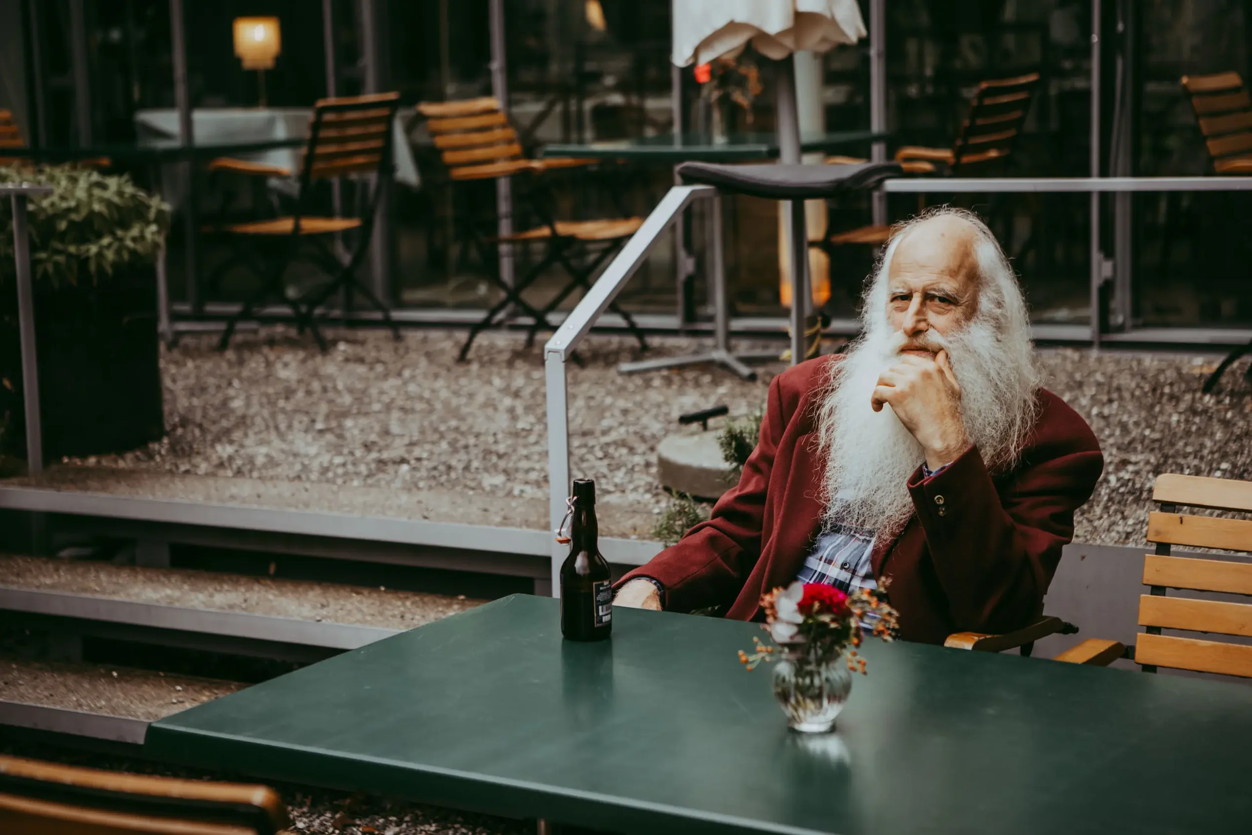 Älterer Mann mit weißem Bart, rote Jacke und Hemd sitzt an einem Tisch in einem Restaurant oder Café, mit einer Flasche Bier vor ihm, im Hintergrund leere Stühle und Tische.