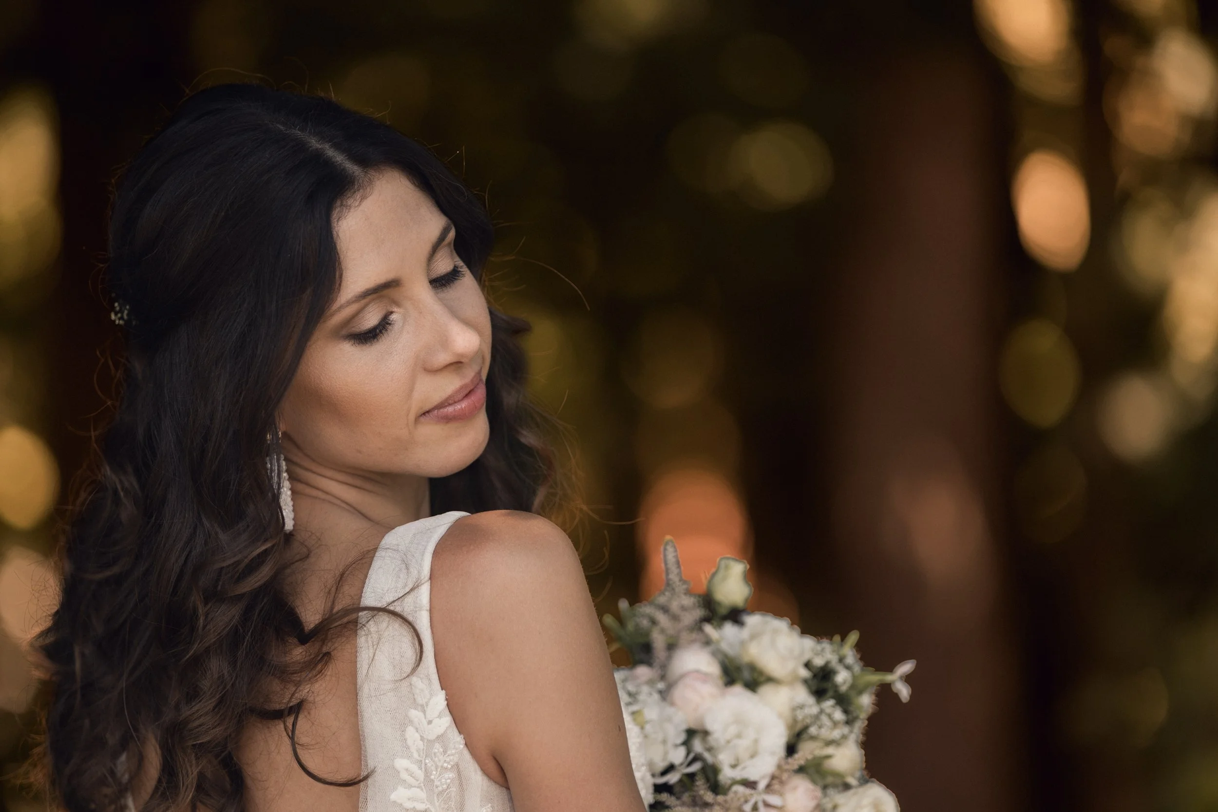 Eine Frau mit dunklen, lockigen Haaren, die auf einer Hochzeit einen Blumenstrauß hält, vor einem Hintergrund mit warmem, bokeh-artigem Licht.
