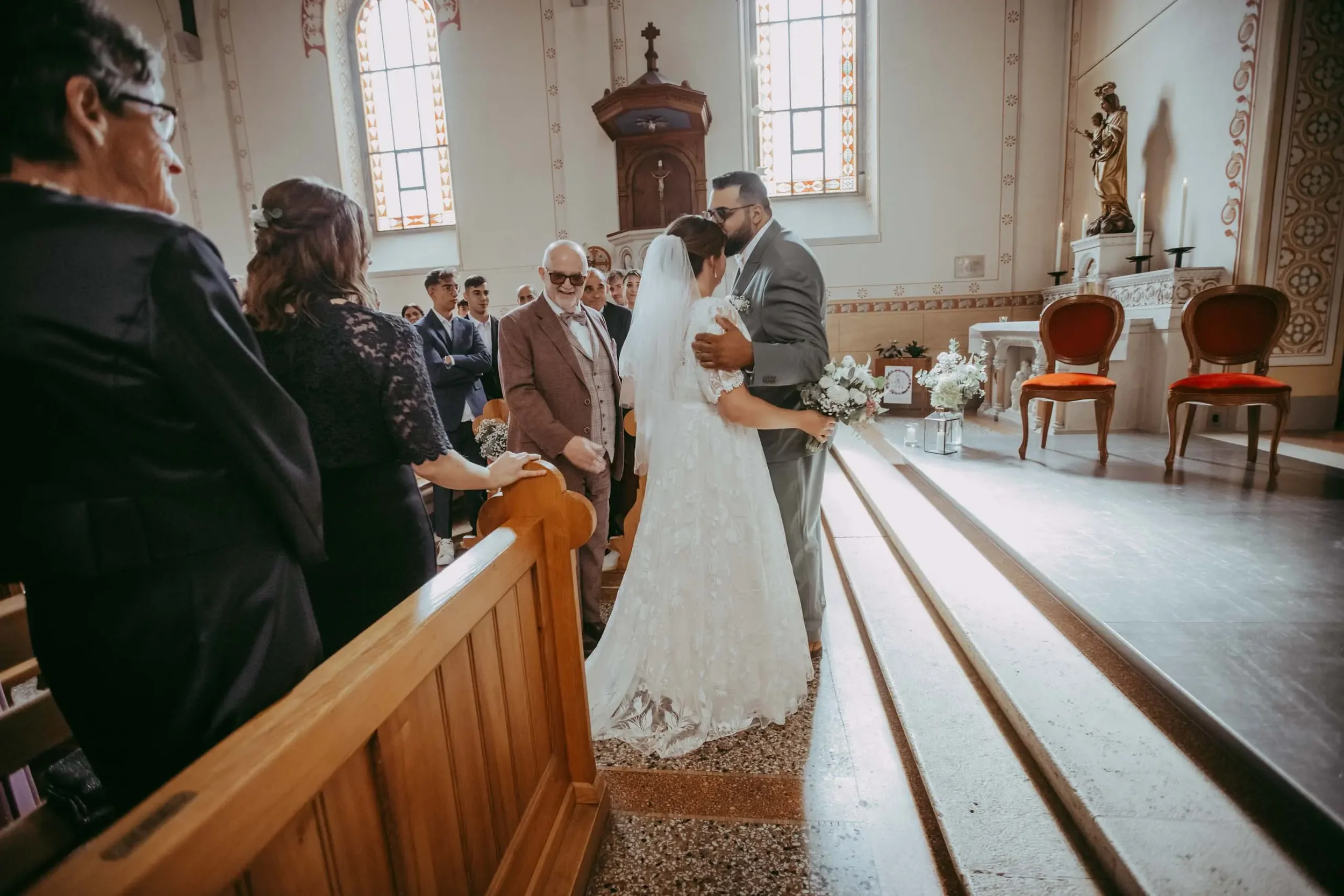 Ein Brautpaar bei einer Hochzeit in einer Kirche, umgeben von Gästen. Sie stehen vor dem Altar, während die Zeremonie stattfindet.