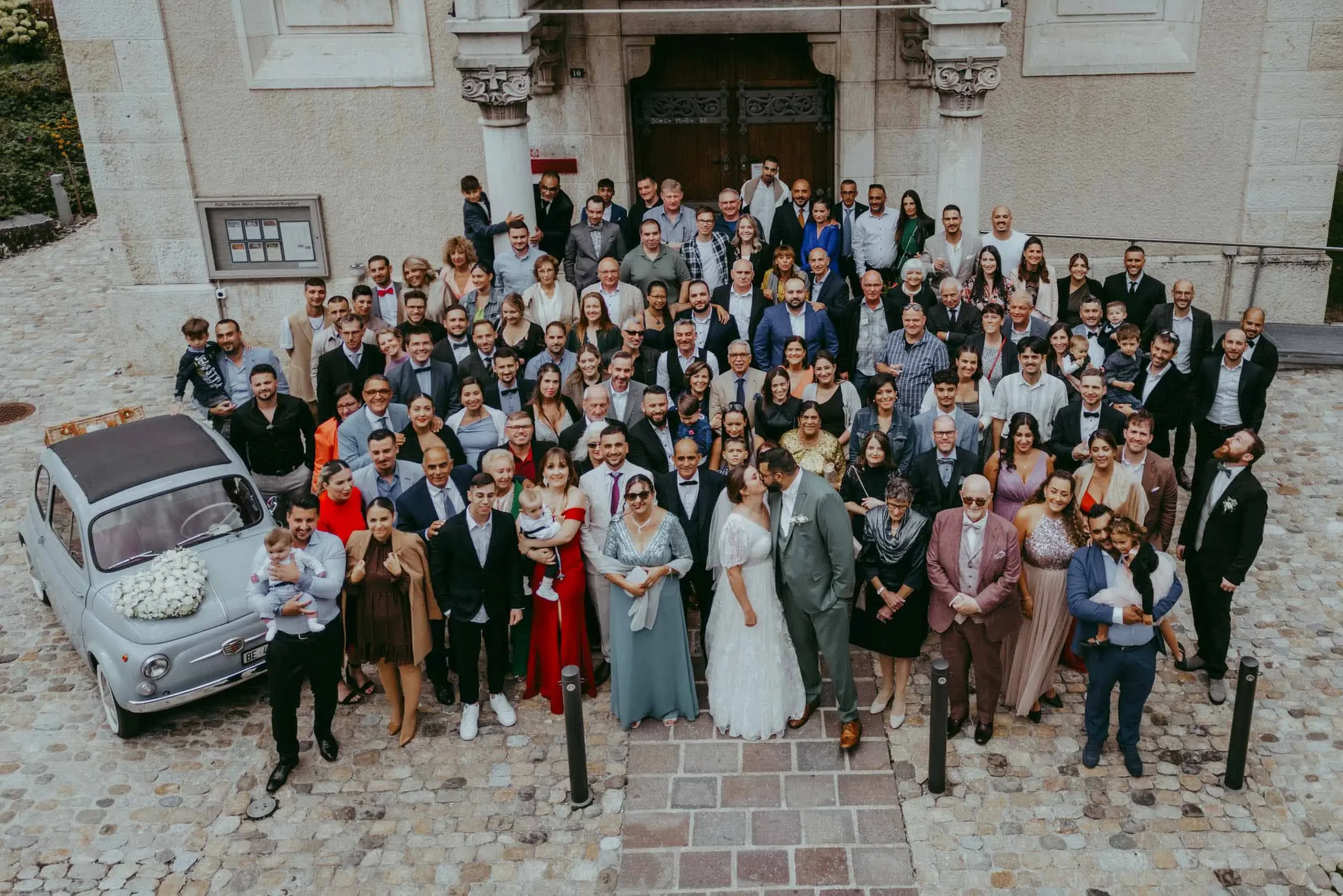 Eine große Gruppe von Menschen vor einer alten Steinaufnahme, vermutlich nach einer Hochzeit, mit Kirchen- oder Schlossgebäude im Hintergrund, inklusive Brautpaar, Familien und Freunden.