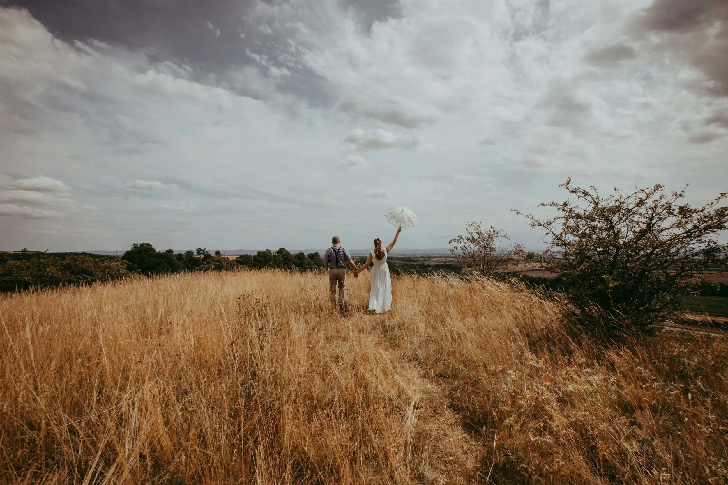 Ein Mann und eine Frau halten Händchen in einem goldenen Feld, während die Frau einen weißen Regenschirm hochhält, mit dunklen Wolken am Himmel im Hintergrund.