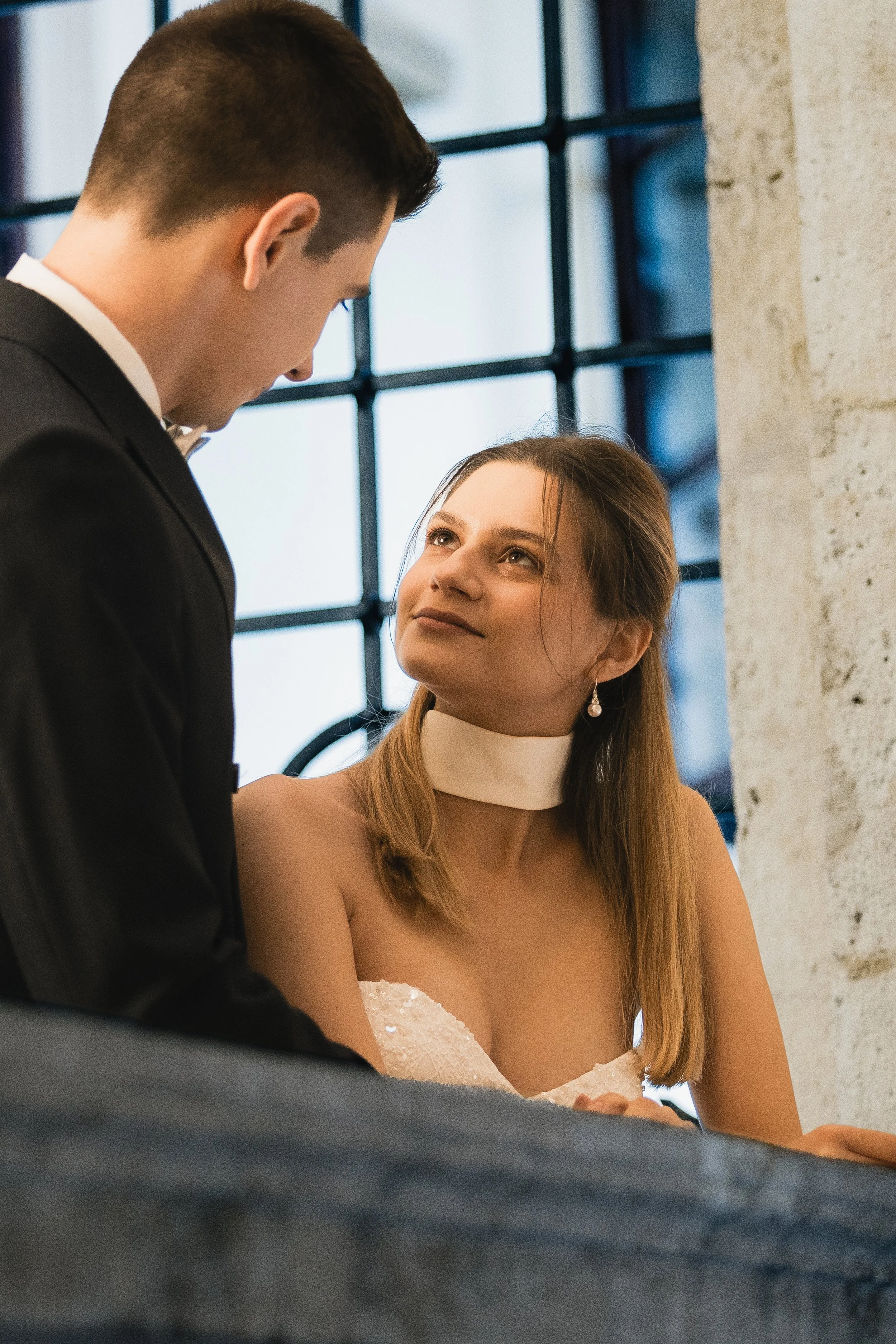 Ein Mann in einem Anzug spricht mit einer Frau in einem weißen Kleid, vermutlich bei einer formellen Veranstaltung oder Hochzeit, in einem Raum mit großen Fenstern und einer Betonwand.