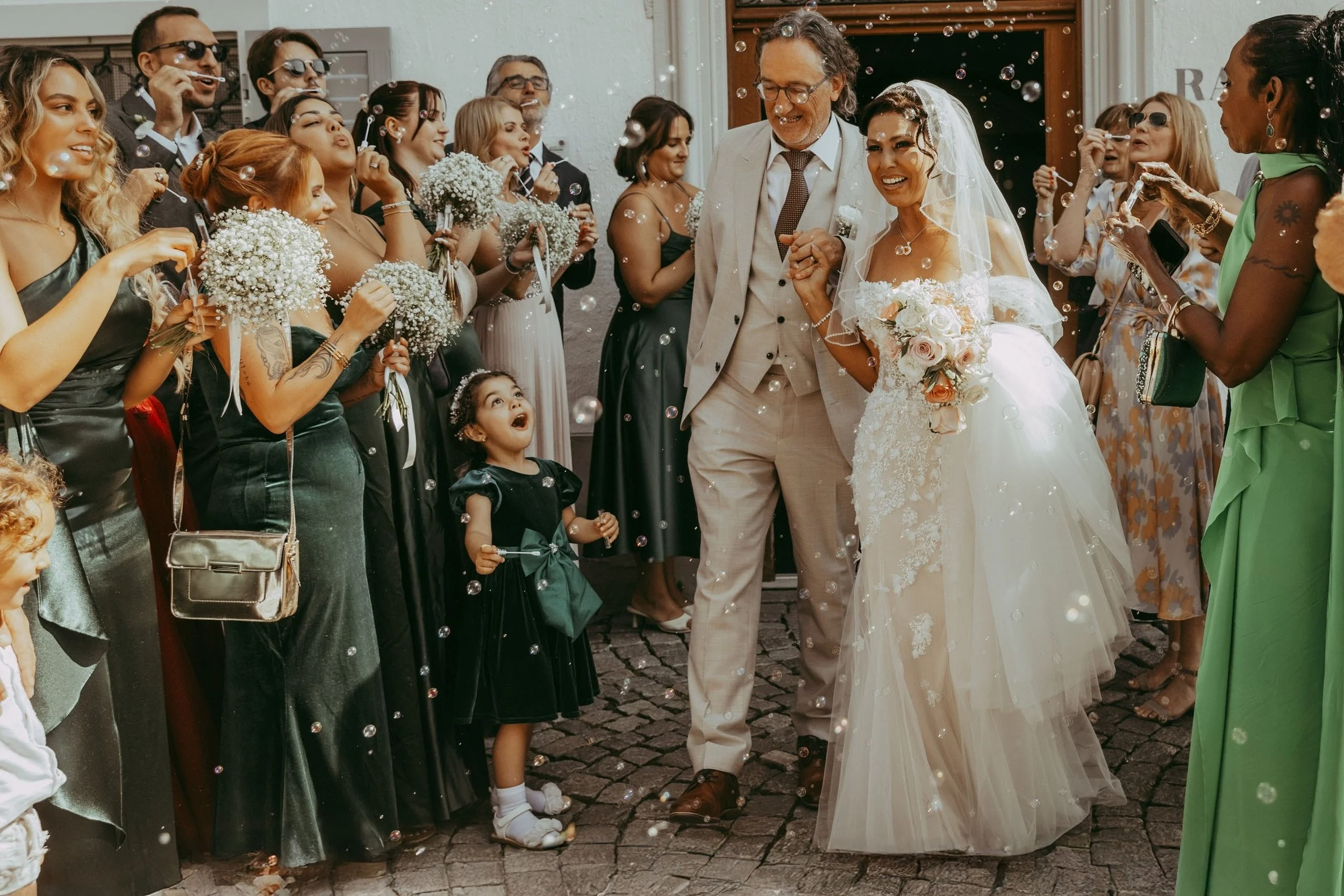 Hochzeit mit Brautpaar, das Händchen haltend, umgeben von Gästen, die Seifenblasen machen und Blumen, alle lächelnd und festlich gekleidet, vor einer weißen Wand.
