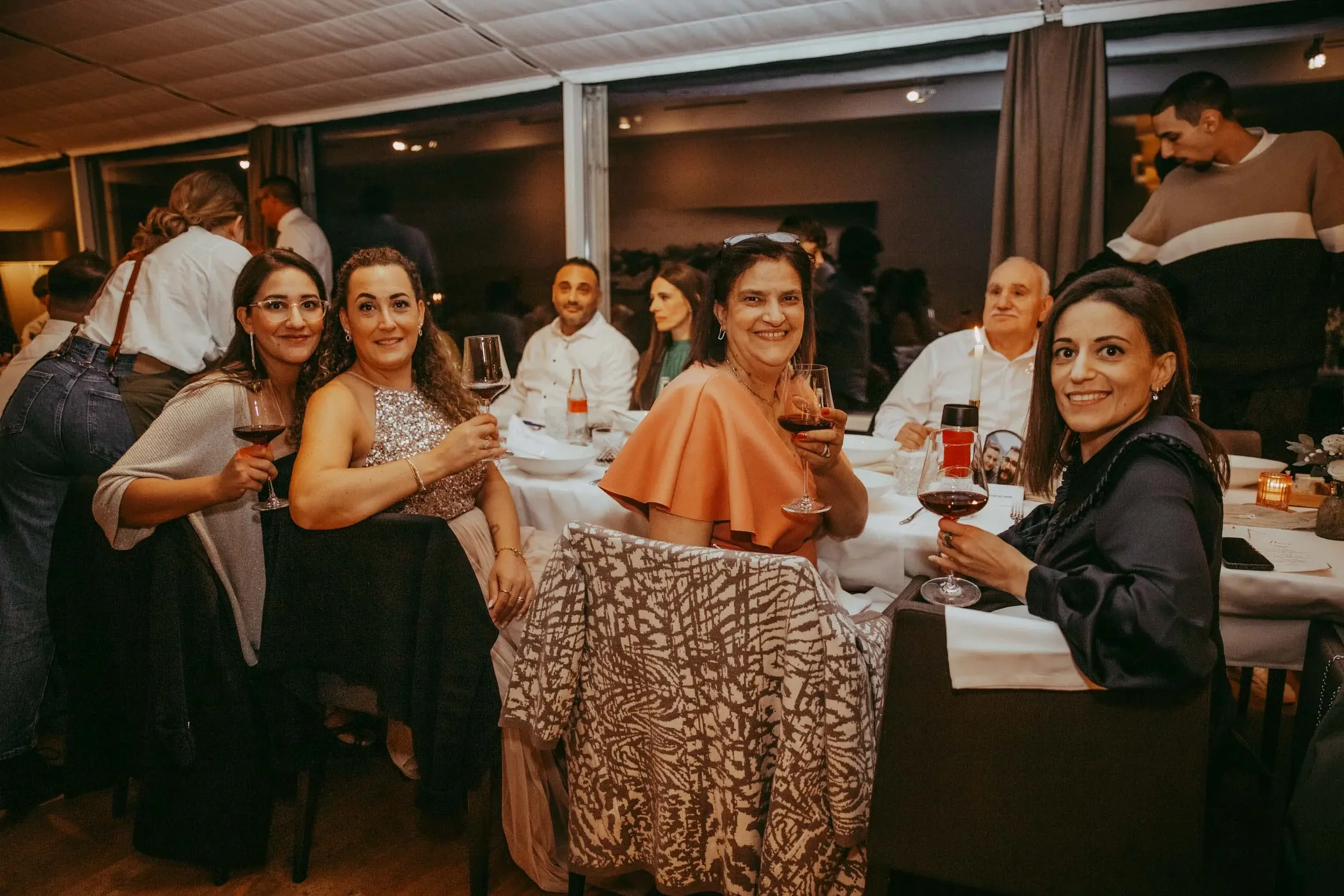 Menschen sitzen an einem festlich gedeckten Tisch, trinken Rotwein, in einem eleganten Restaurant bei Abenddämmerung, lachend und kommunikativ, im Hintergrund weitere Gäste.