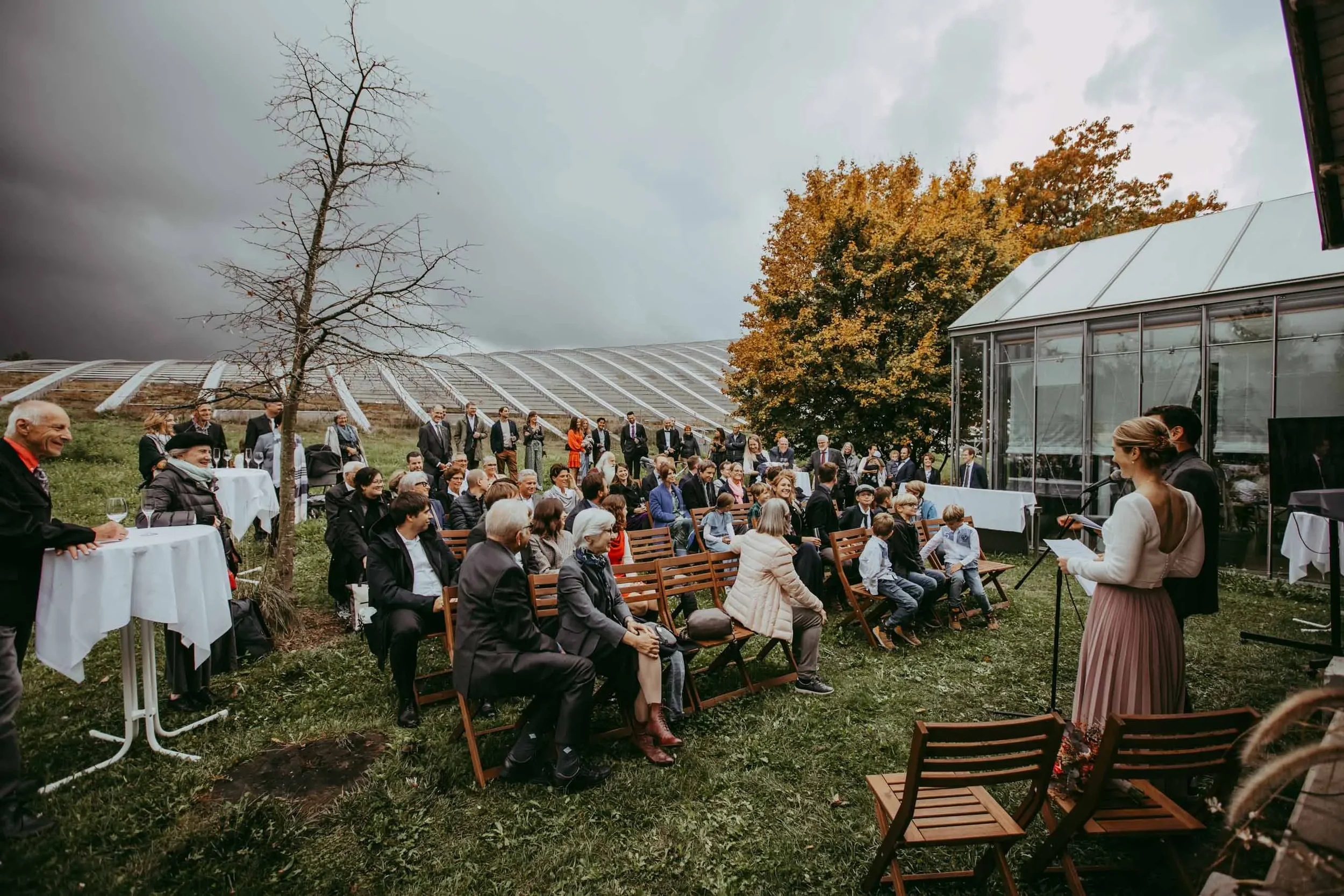 Eine Gruppe von Menschen bei einer outdoor Veranstaltung, einige sitzen auf Stühlen, andere stehen. Es befindet sich eine Rednerin an einem Mikrofon rechts im Bild, im Hintergrund sind Bäume und ein Gewächshaus. Das Wetter wirkt wechselhaft mit dunkl