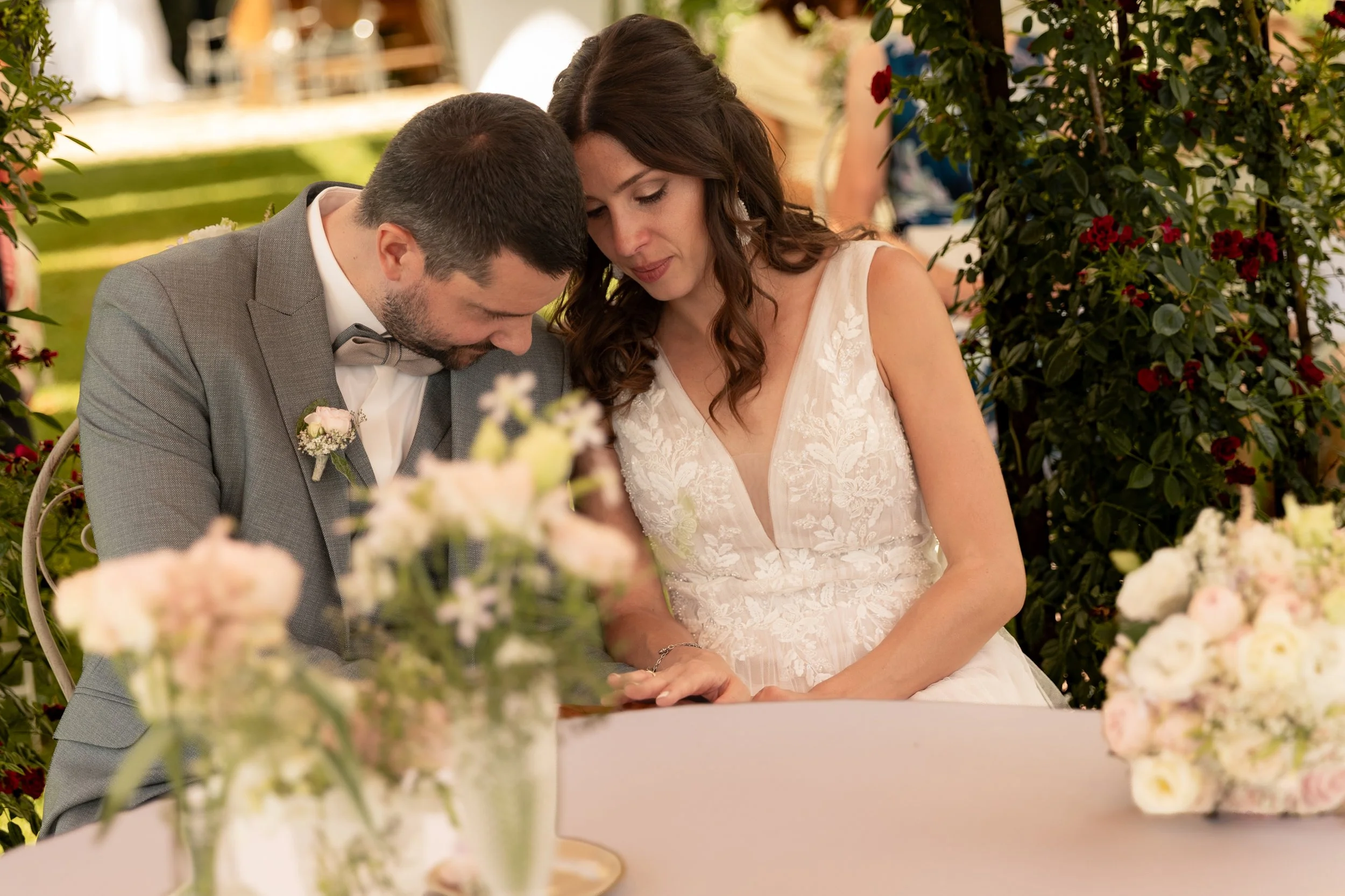 Ein Brautpaar bei einer Hochzeit, beide sitzen eng beieinander und scheinen in einem emotionalen Moment zu sein, umgeben von Blumen im Freien.