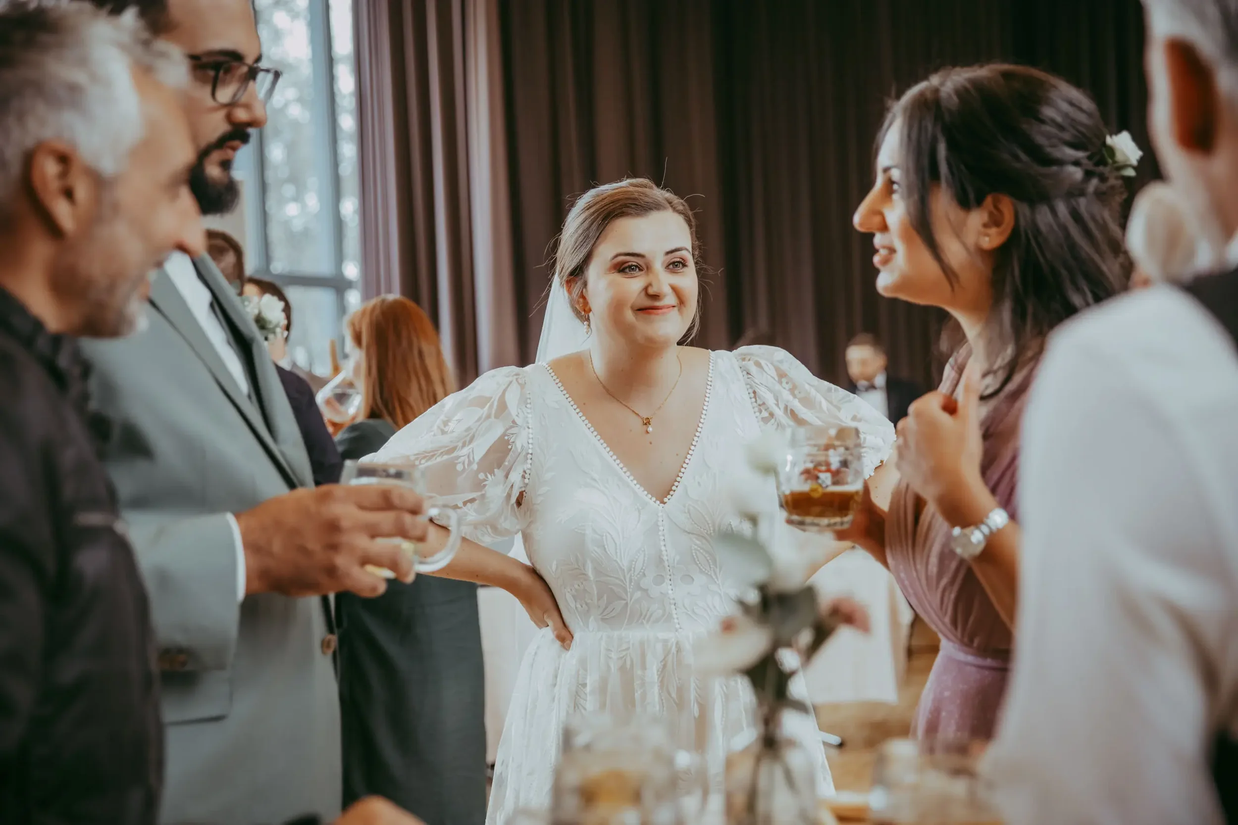 Hochzeitspaar, Frau in Weiß, und Gäste, Männer in Anzügen im Gespräch bei einer Feier in einem Raum mit dunklen Vorhängen.