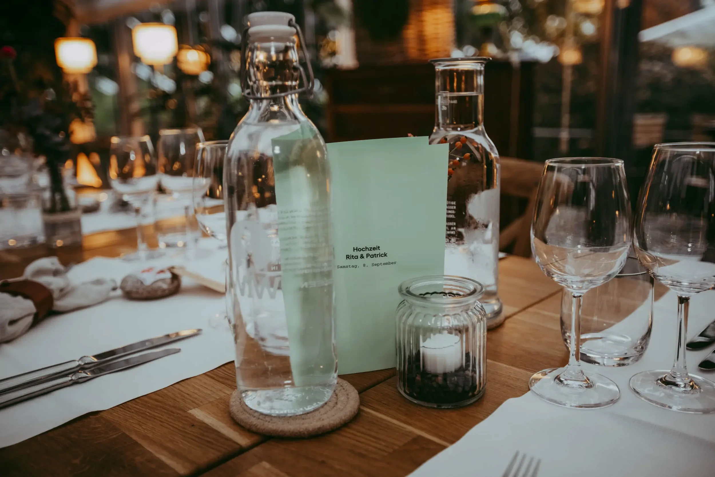 Tischdekoration bei einer Hochzeit mit Wasser, Gläser, Kerzen und einem Hochzeitsmenü in der Mitte, im Hintergrund eine warme Beleuchtung.