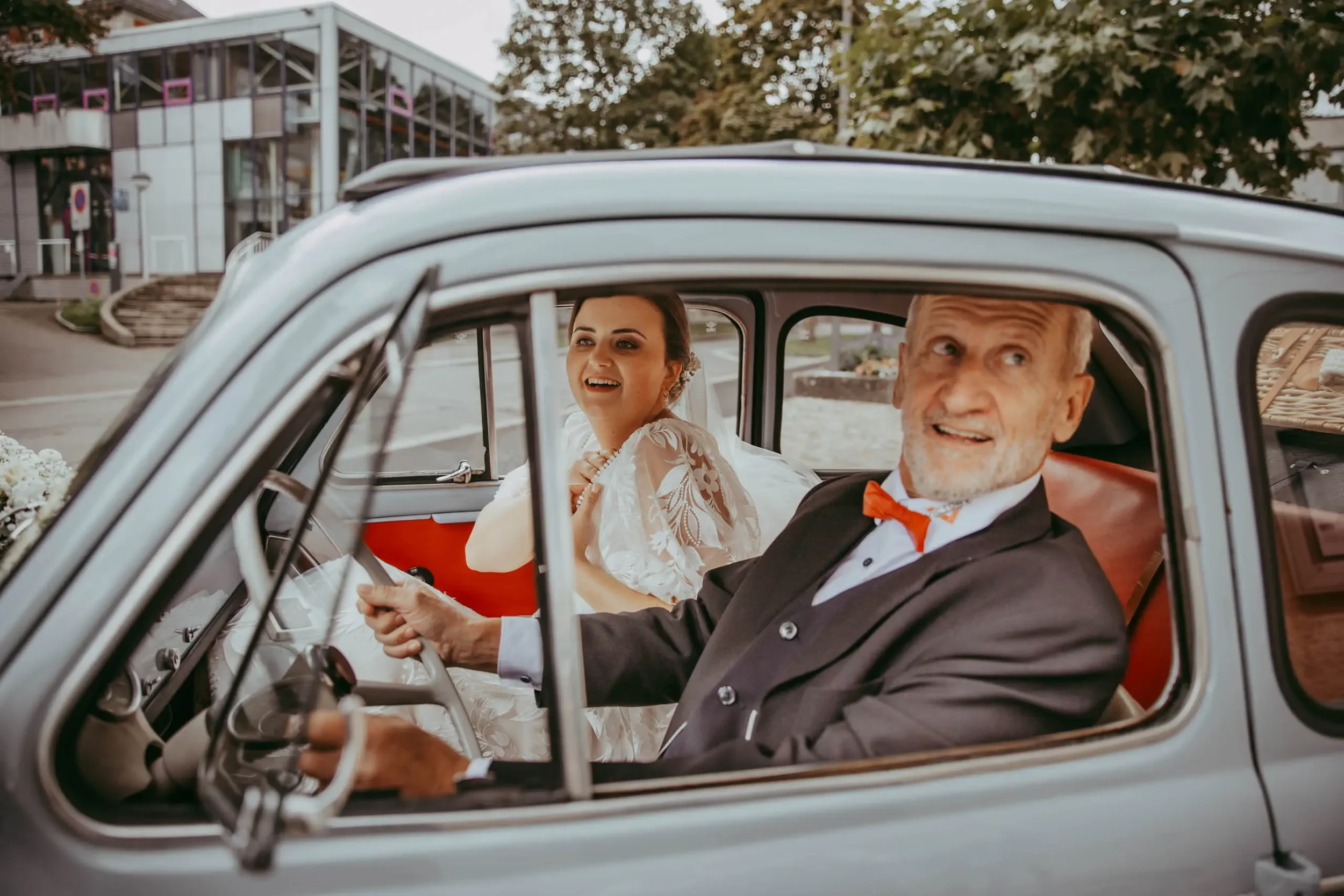 Ein junges Brautpaar sitzt in einem silbernen Oldtimer, der während ihrer Hochzeit als Fahrzeug dient. Die Braut trägt ein weißes Hochzeitskleid mit Stickereien, während der Bräutigam einen schwarzen Anzug mit roter Fliege trägt. Das Paar lächelt und