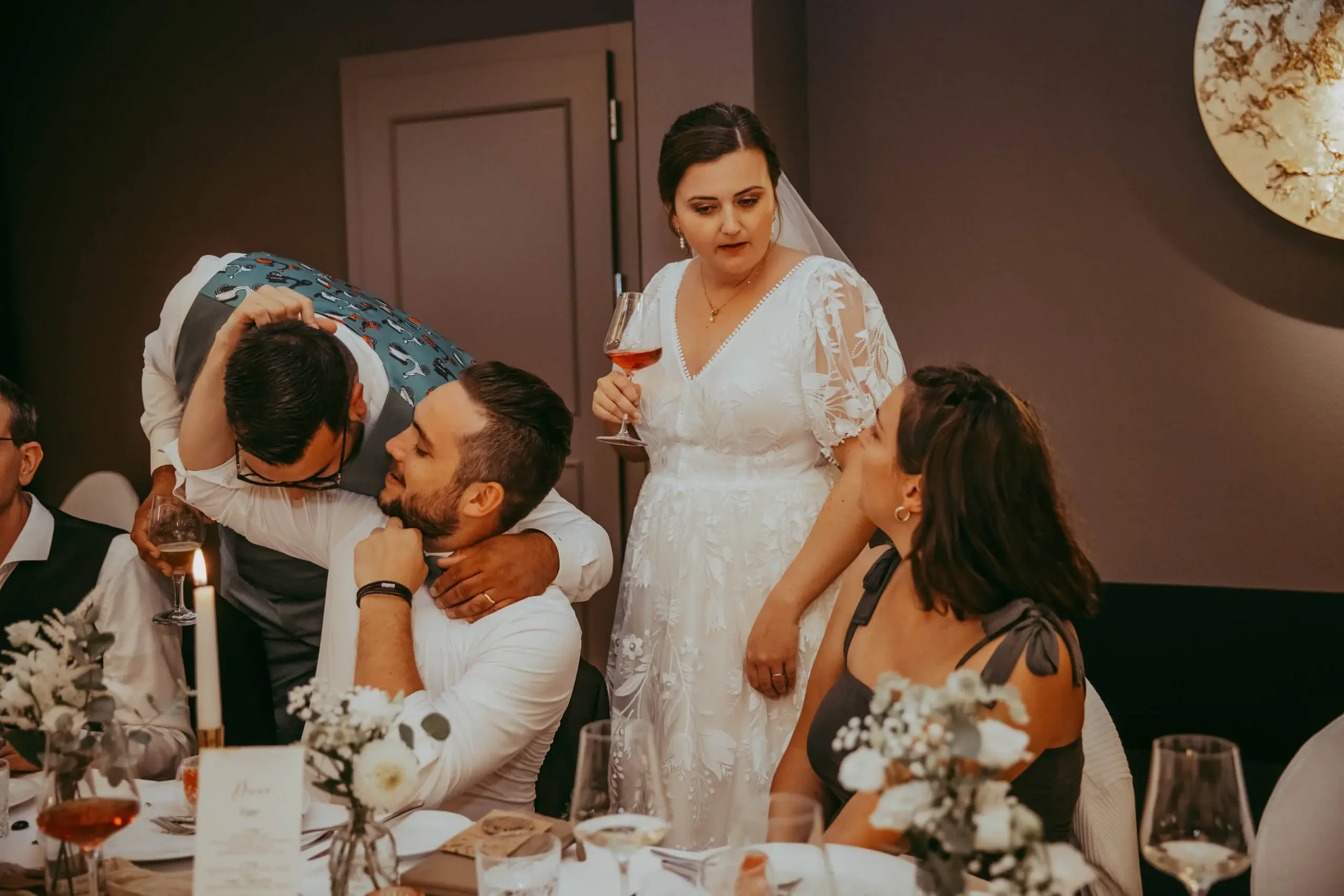 Eine Braut trägt ein weißes Hochzeithand in einer eleganten Hochzeitsszene, umgeben von Gästen, mit einem Glas Wein in der Hand, während andere Gäste an einem festlich gedeckten Tisch sitzen.
