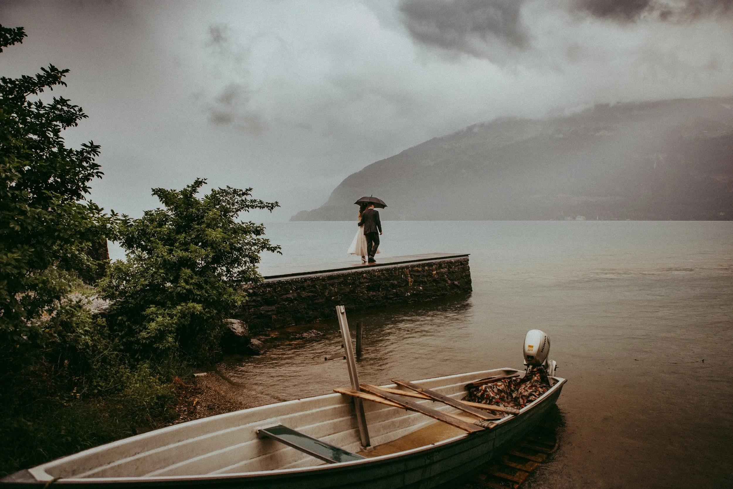 Ein Paar läuft an einem Ufer bei Regen, hält sich unter einem Regenschirm, im Vordergrund Boote am Ufer, im Hintergrund dunkler Himmel mit Wolken und Bergen.