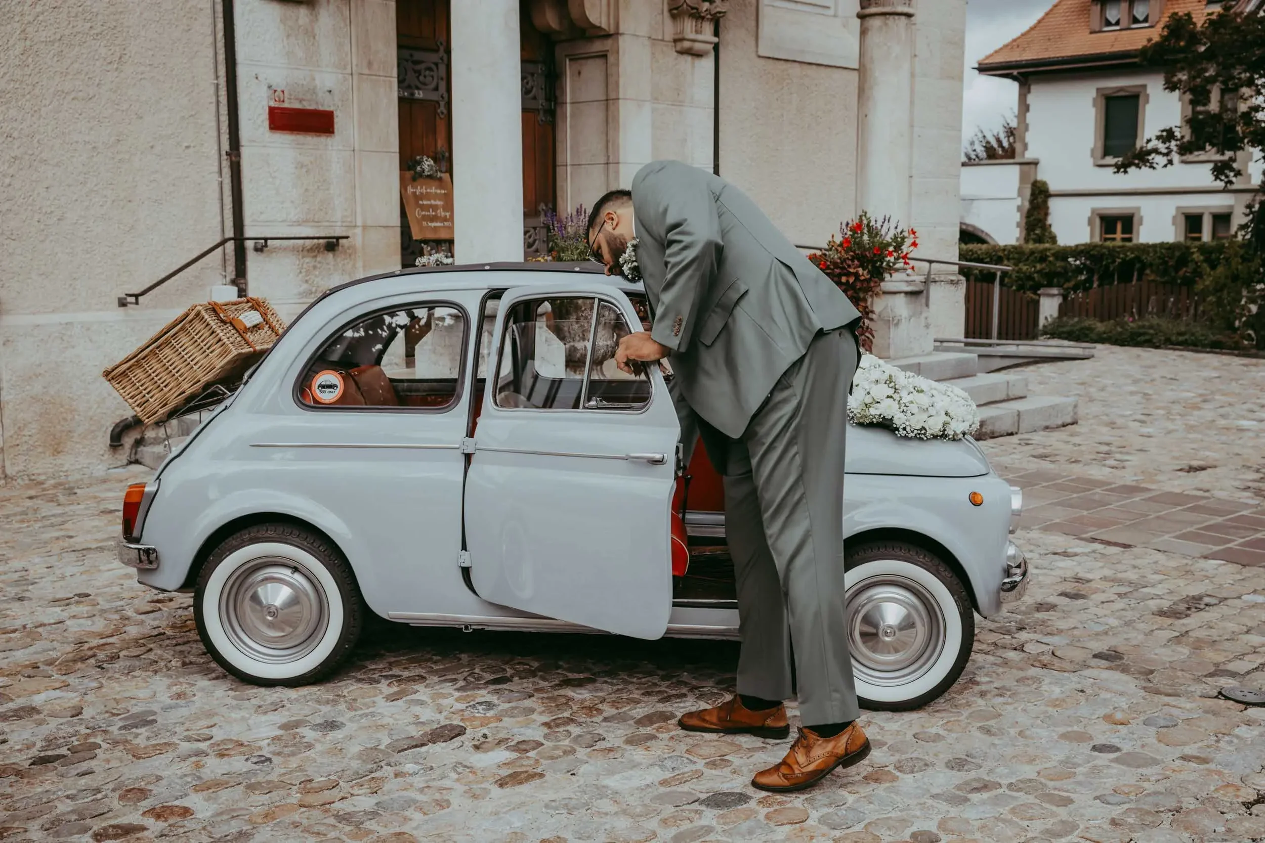 Ein Mann im grauen Anzug steigt in einen kleinen, silbernen Oldtimer mit offener Tür und einem Korb auf dem Dach, vor einem historischen Gebäude aus Stein. Es sind Blumen und Pflanzen im Hintergrund sichtbar.