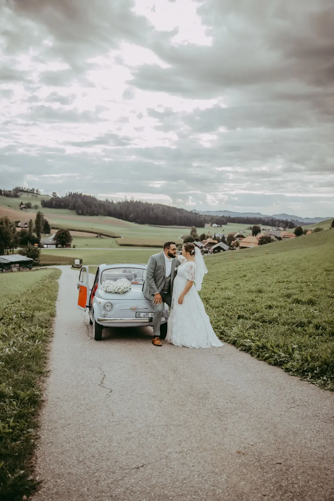 Ein Brautpaar in Hochzeitskleidung steht auf einer Straße mit einem kleinen blauen Auto, im Hintergrund grüne Hügel und eine ländliche Landschaft, bewölkter Himmel.