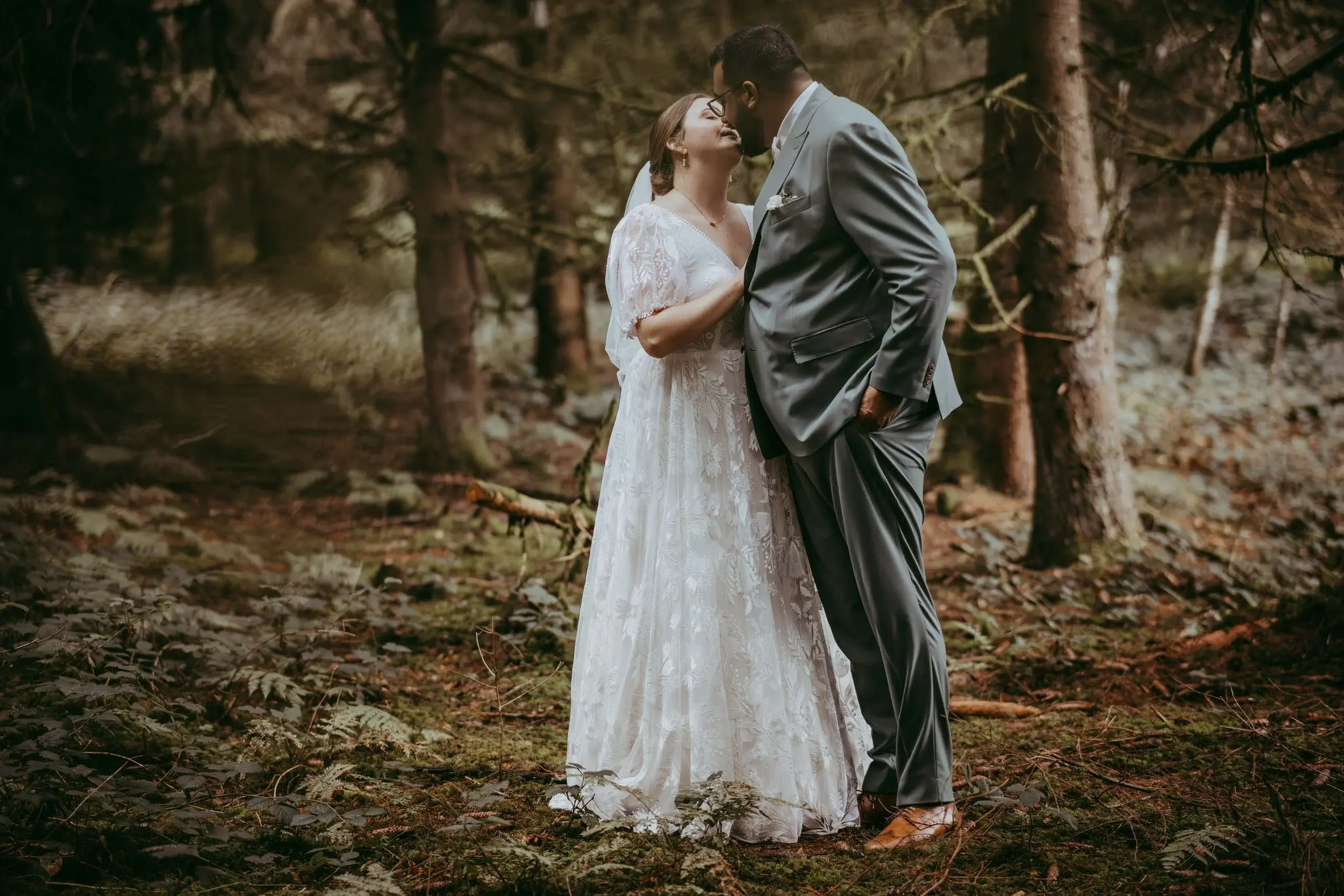 Ein Brautpaar in formeller Kleidung, die sich in einem Wald befinden, berühren sich mit den Nasen. Die Frau trägt ein weißes Hochzeitskleid, der Mann einen grauen Anzug.
