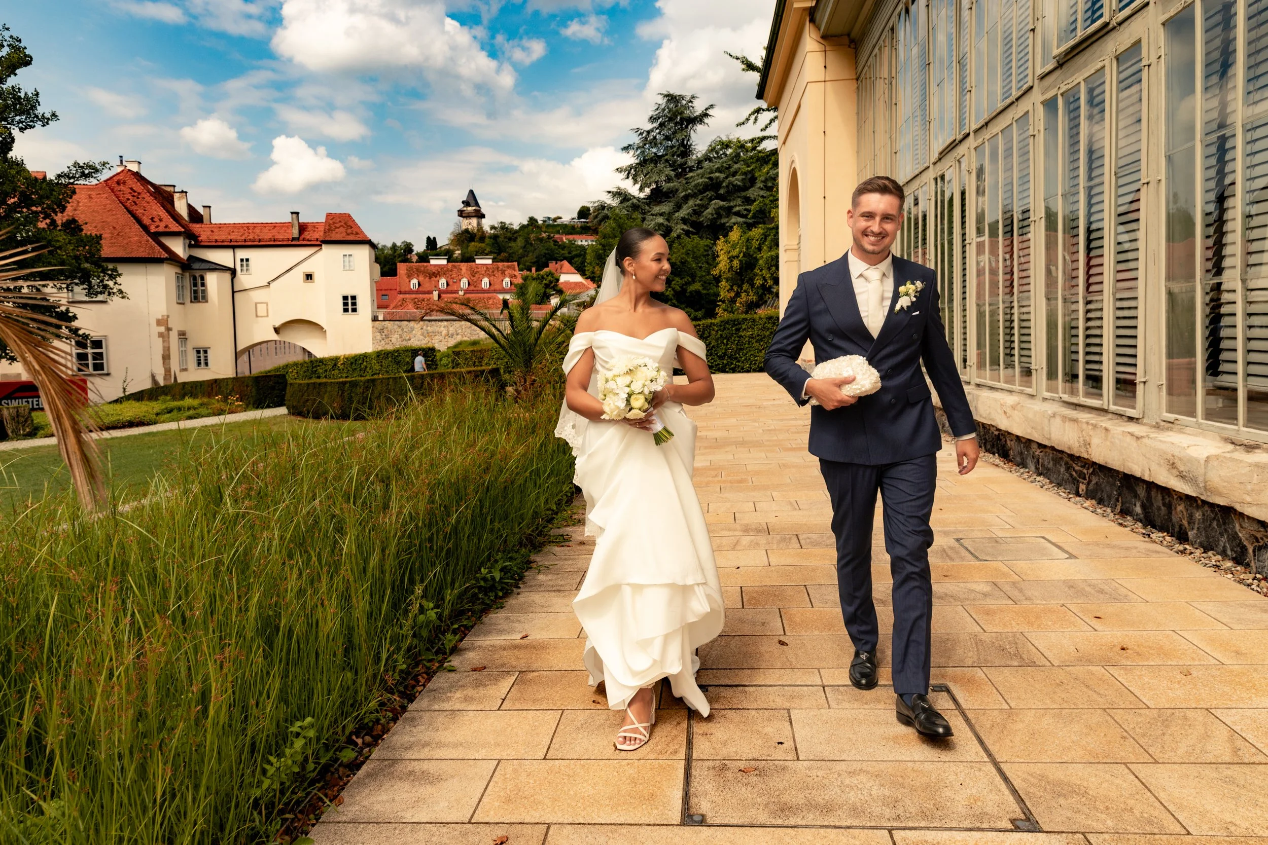 Ein Brautpaar bei der Hochzeit, die Braut trägt ein weißes Hochzeitskleid und hält einen Blumenstrauß, der Bräutigam trägt einen dunklen Anzug und hält ein Geschenk, sie gehen auf einem gepflasterten Weg am Gebäude entlang.