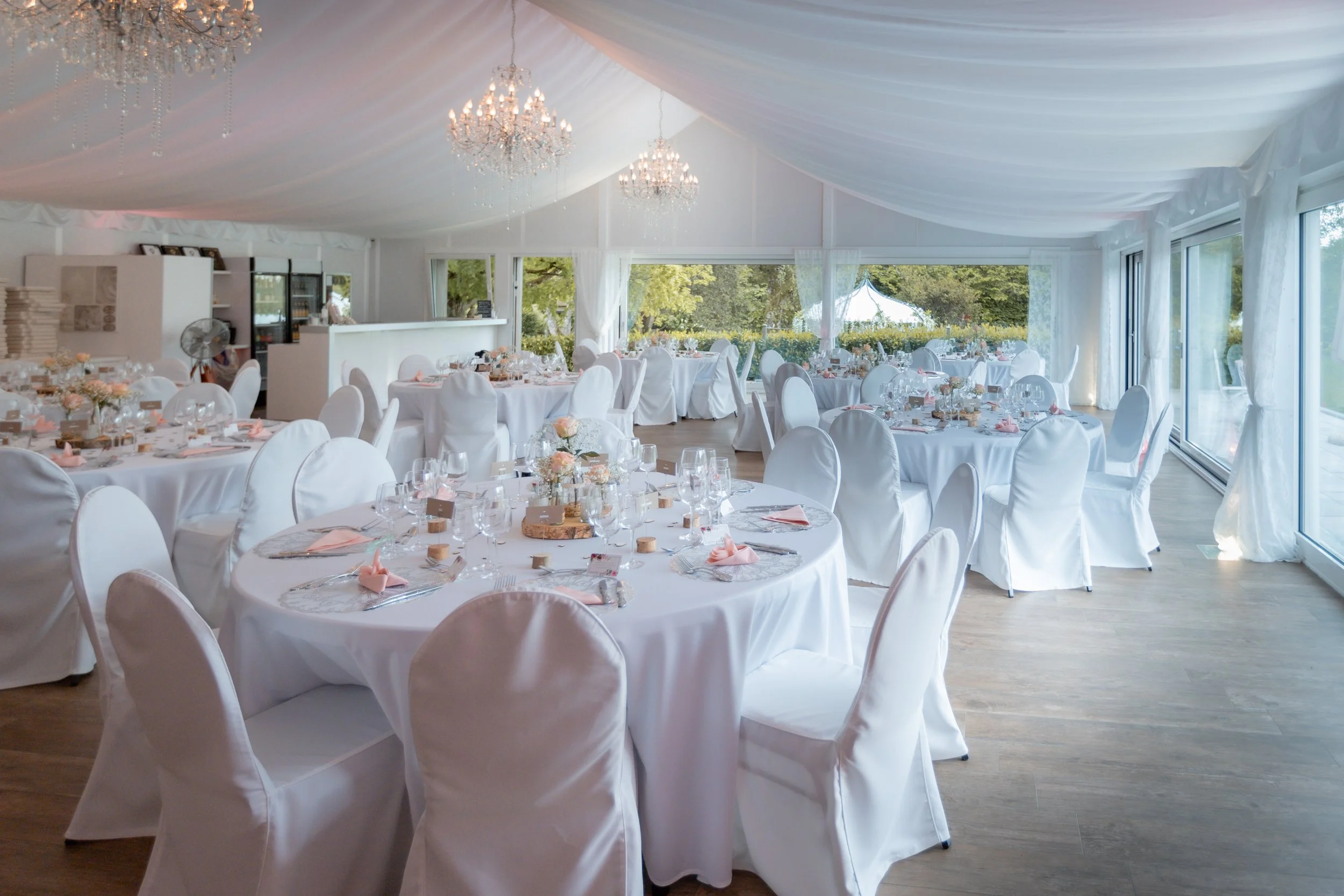 Ein eleganter festlich gedeckter Saal mit runden Tischen, weißen Stuhlhüllen, Blumentischdekorationen und Kristalllüstern, die an der weiß gedeckten Decke hängen.