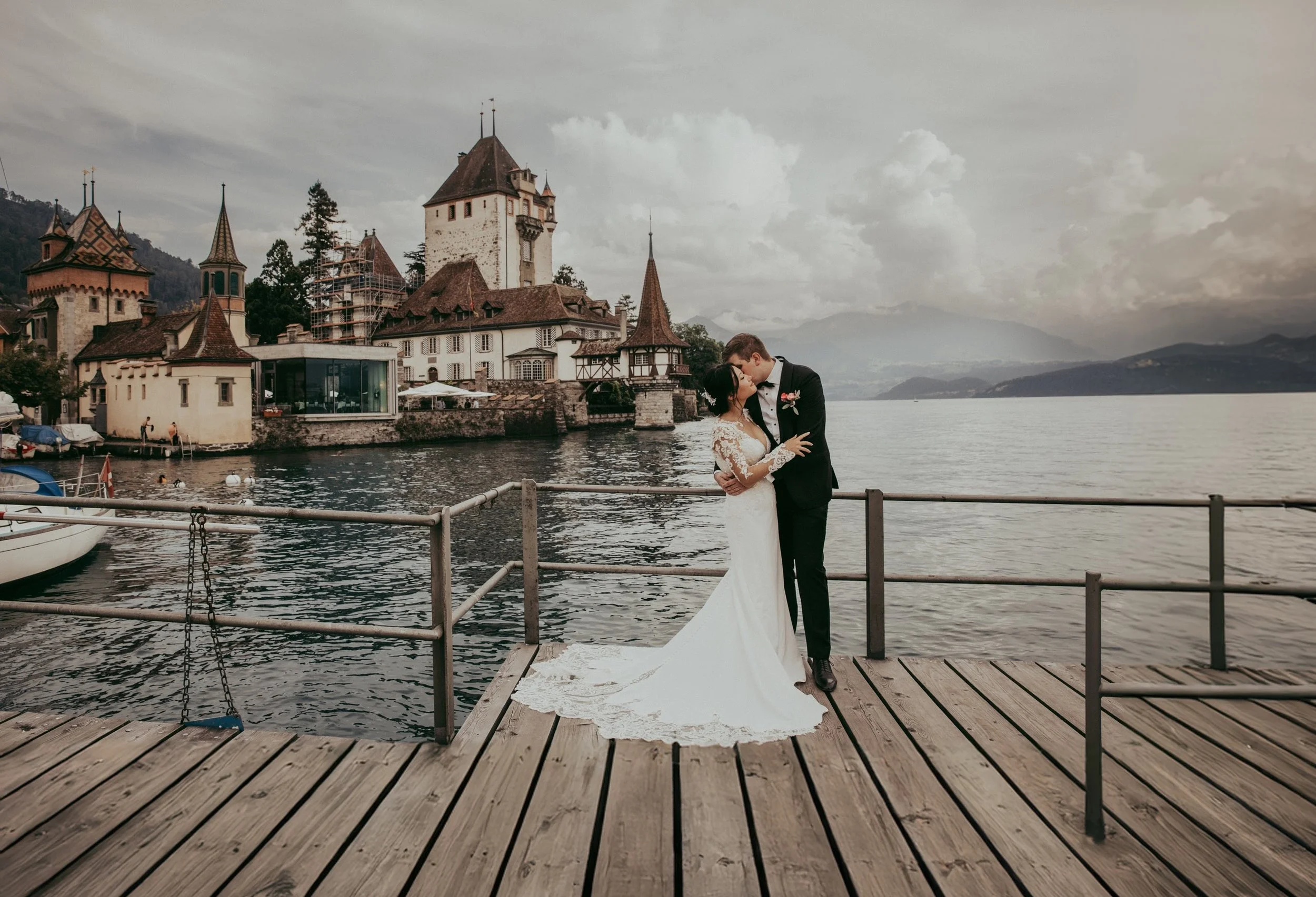 Ein Brautpaar, das sich auf einem Steg am See küsst, mit mittelalterlicher Architektur im Hintergrund, bewölktem Himmel und Wasser